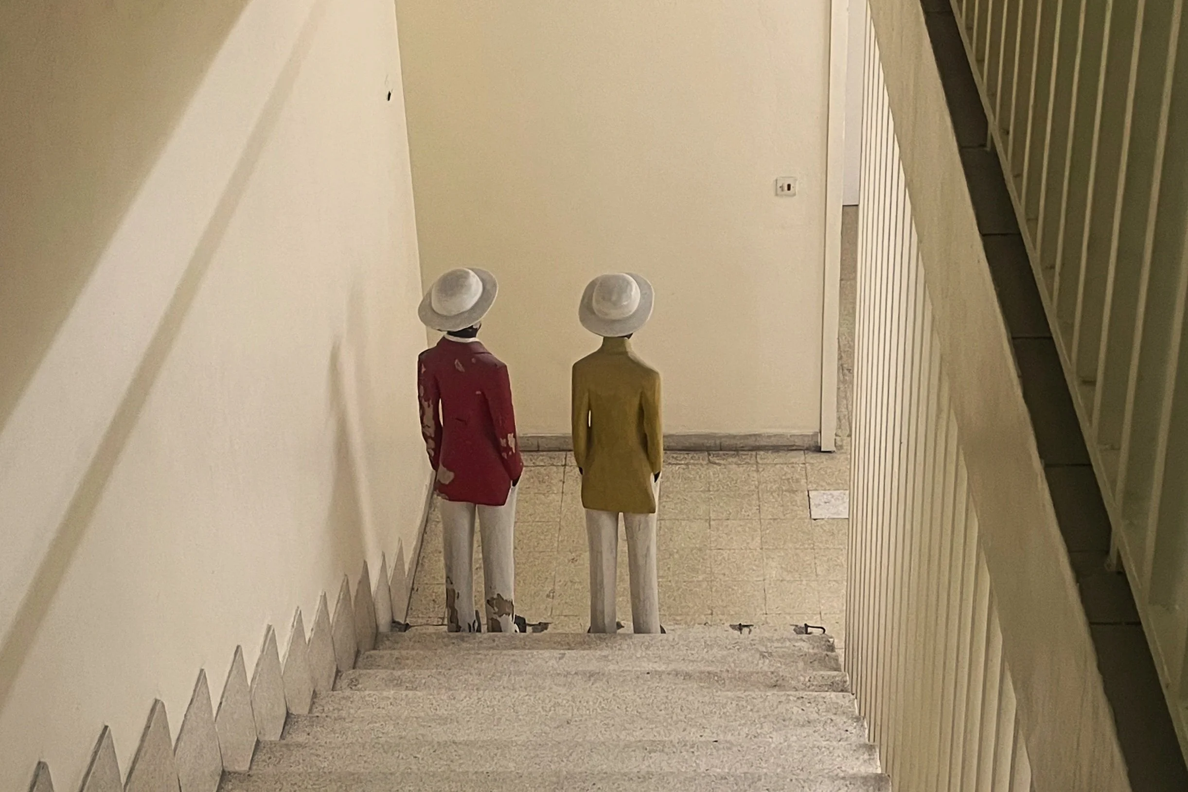 Two mannequins wearing fashionable outfits and hats, positioned at the bottom of a staircase in an indoor space.