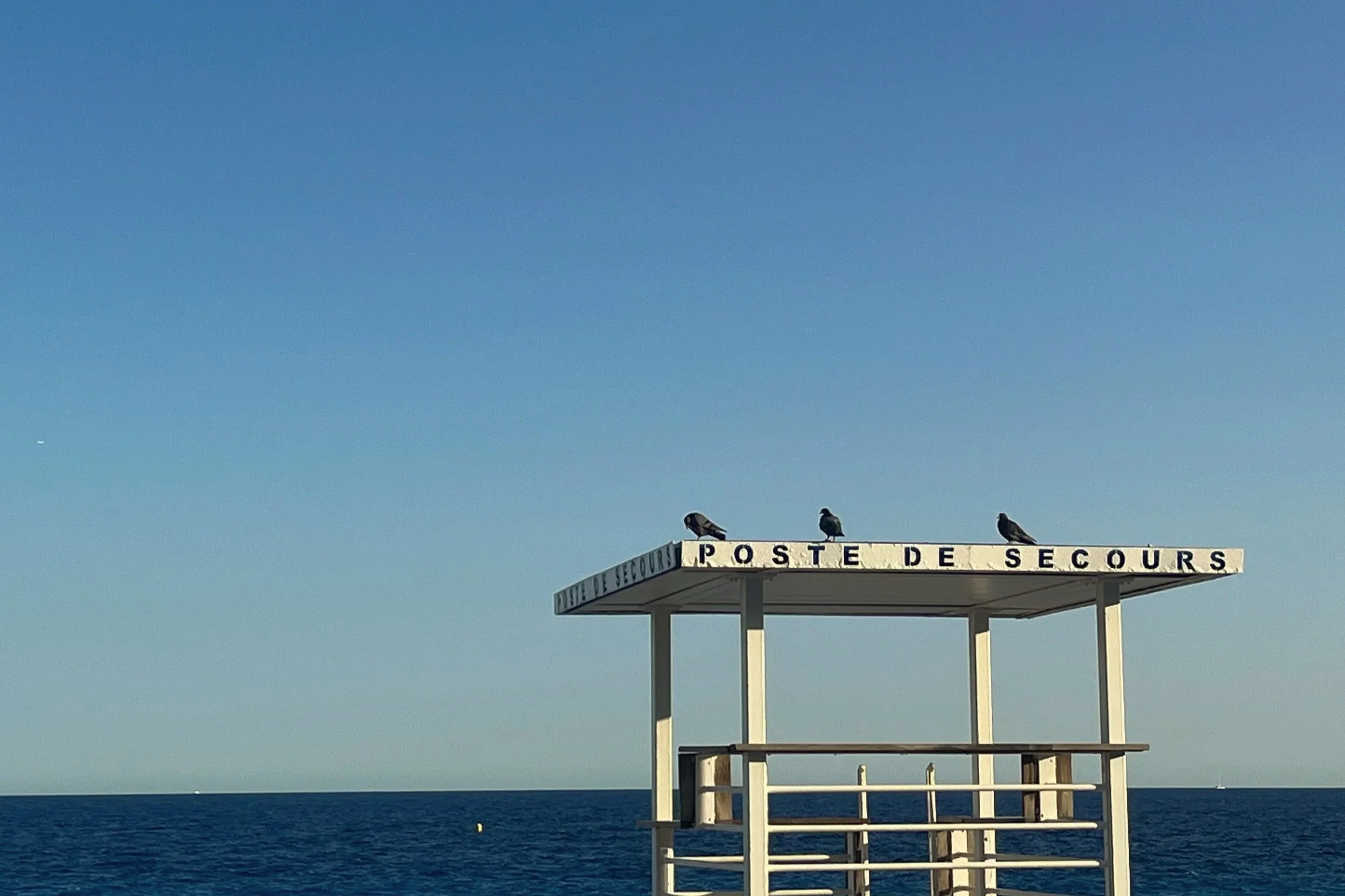 A lifeguard station with the sign 'Poste de Secours' on the roof, three birds perched on top, against a background of the blue sea and sky.