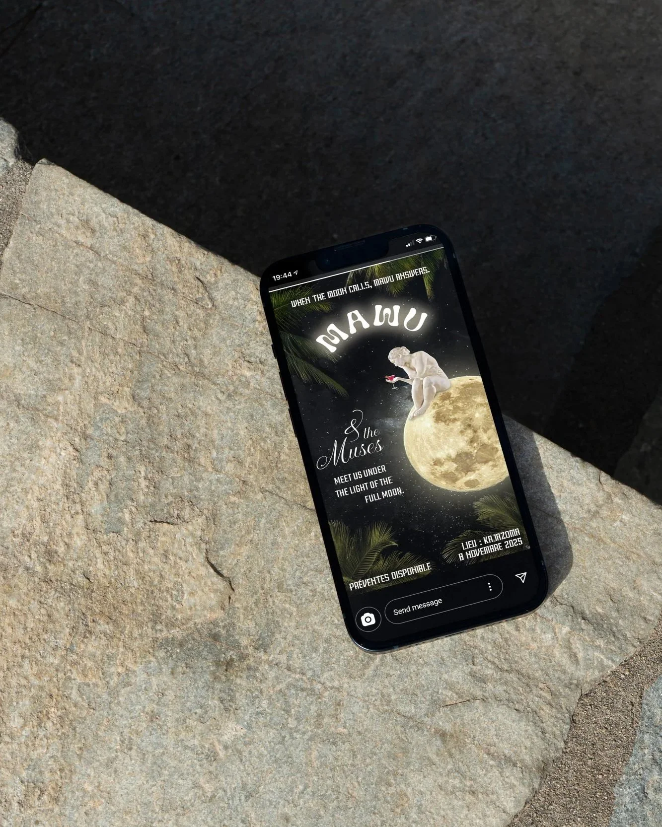 A smartphone displaying a promotional poster for an event called 'Mawu & the Muses' on a stone surface outdoors. The poster features a moon, a classical sculpture of a woman, palm leaves, and event details.