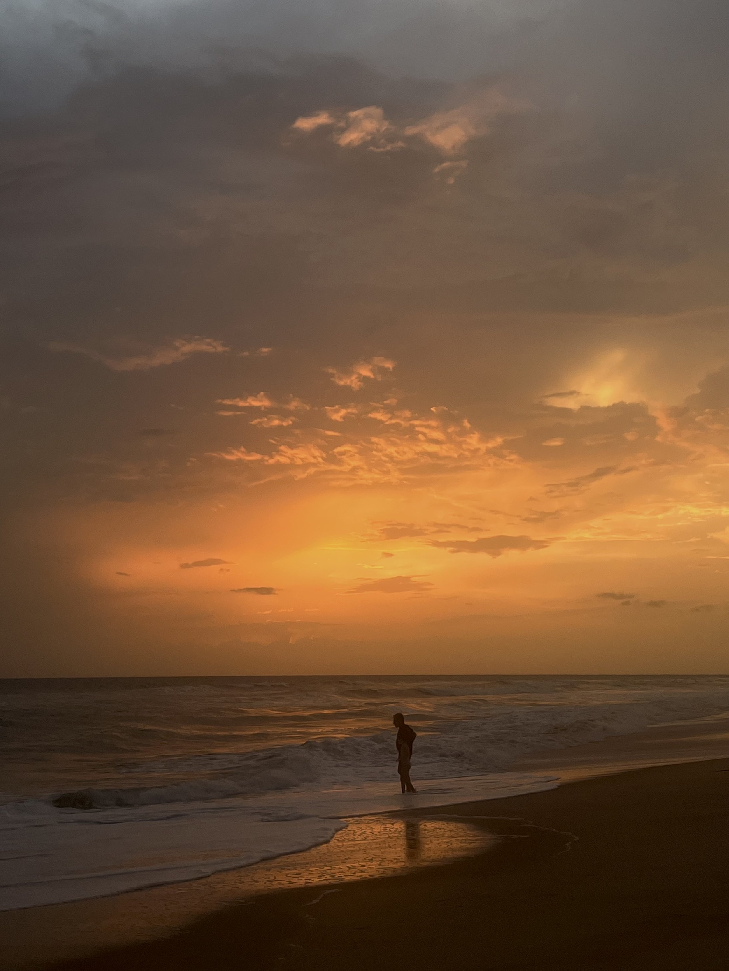 A person standing alone on the shoreline of a beach during sunset, with a colorful sky and calm waves.