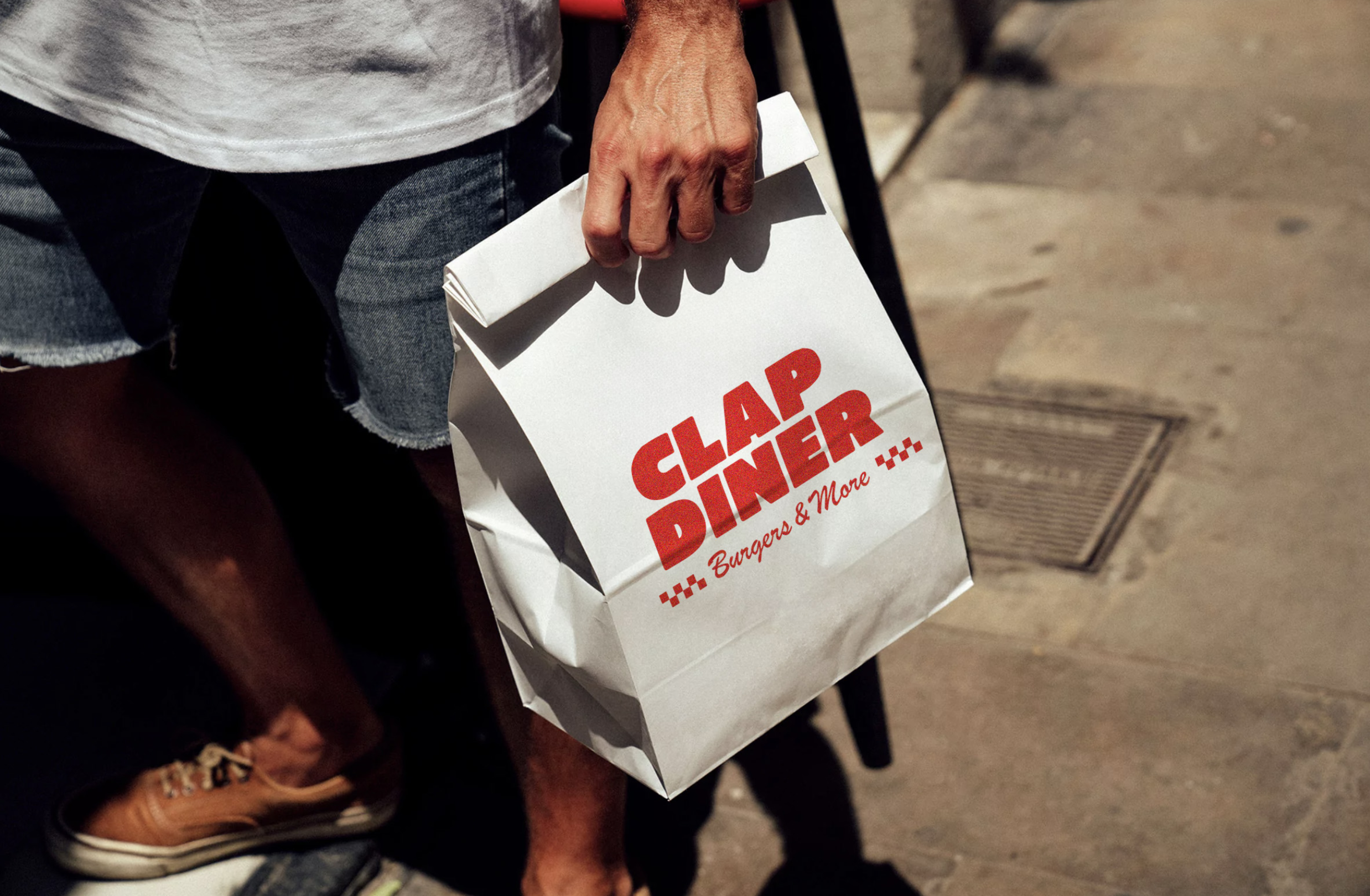 Person holding a white paper bag with the logo 'CLAP DINER' in red and the tagline 'Burgers & More...' in red cursive, standing on a sidewalk.