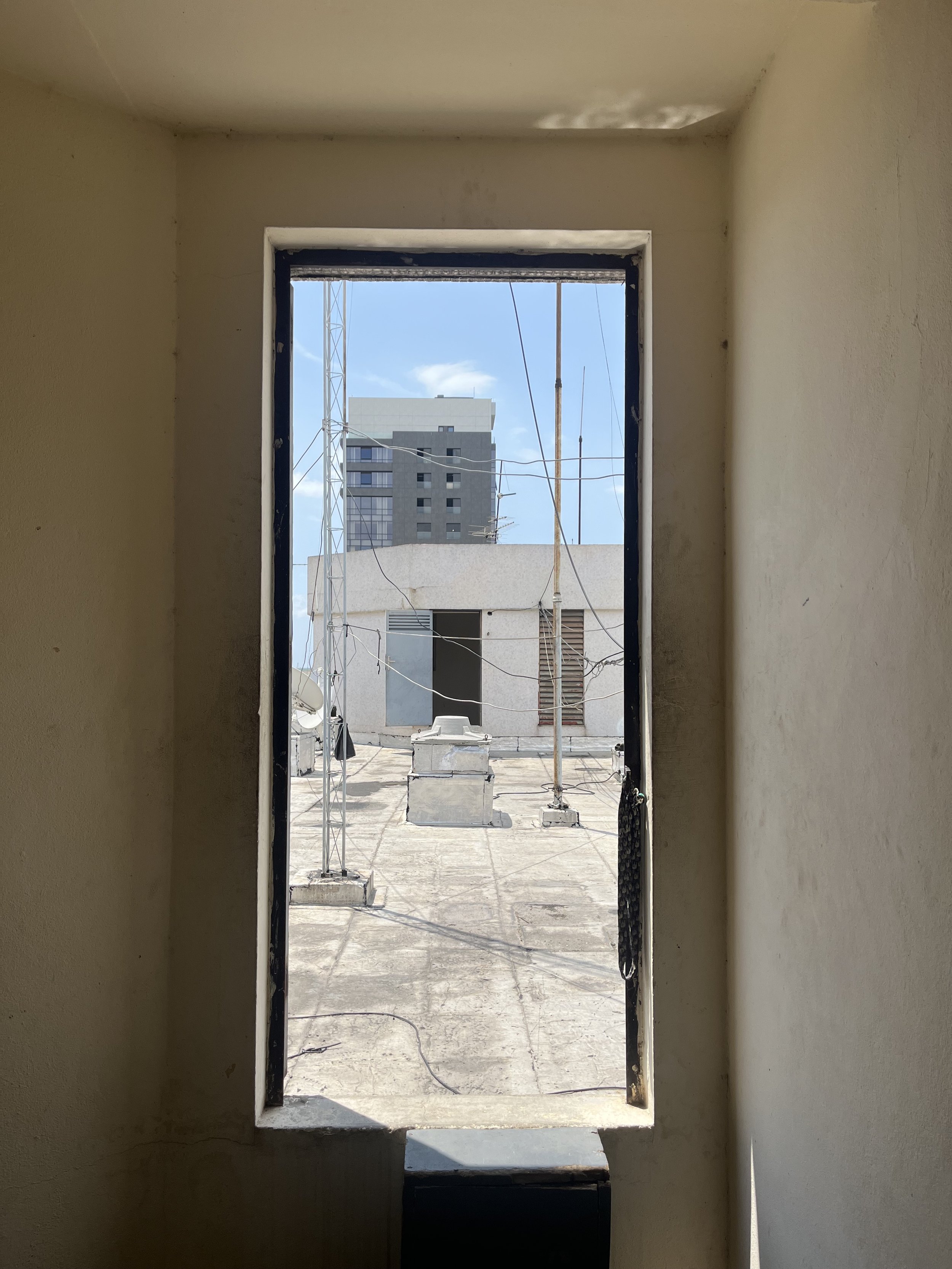 View from inside a small room looking out through a rectangular window at a building rooftop with antennas and wires, and taller buildings in the distance under a blue sky with some clouds.