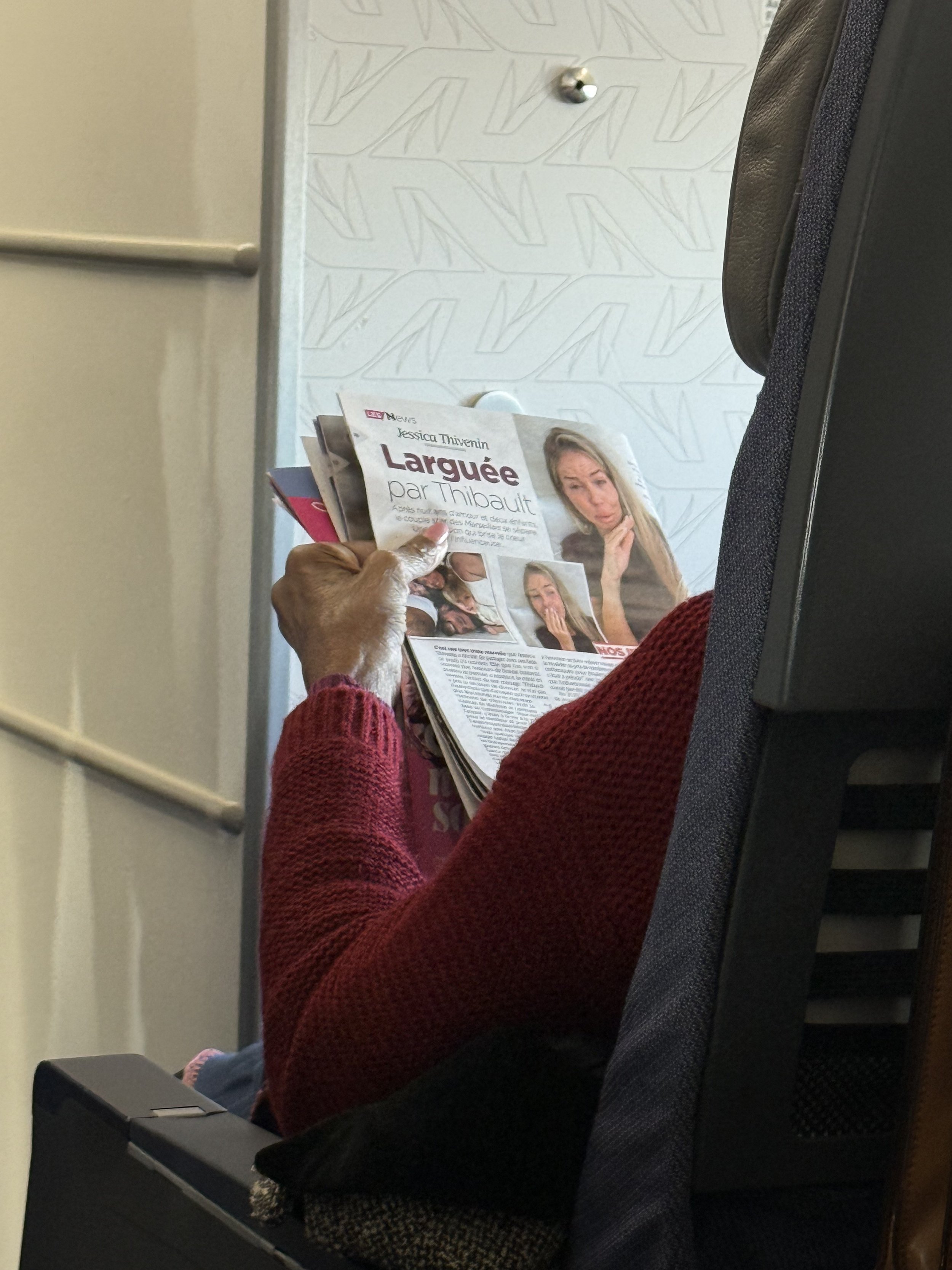 A person with dark skin wearing a red sweater, sitting in an airplane seat, reading a newspaper with a headline in French. The person is holding the newspaper and their face is not visible.