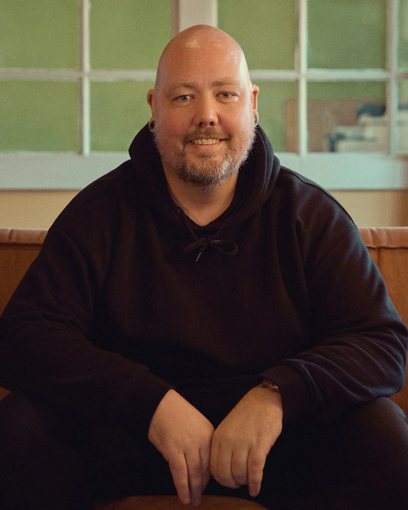 En mann med kort skjegg, som smiler, sitter på en benk innendørs.