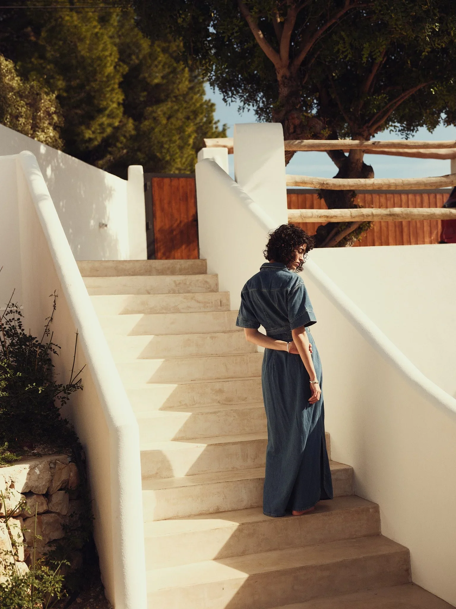 En kvinne i denimklær går ned en utendørs trapp av stein, omgitt av hvite vegger og en stor tre med grønne blader i bakgrunnen. Solskinn skaper skygger på trappen.
