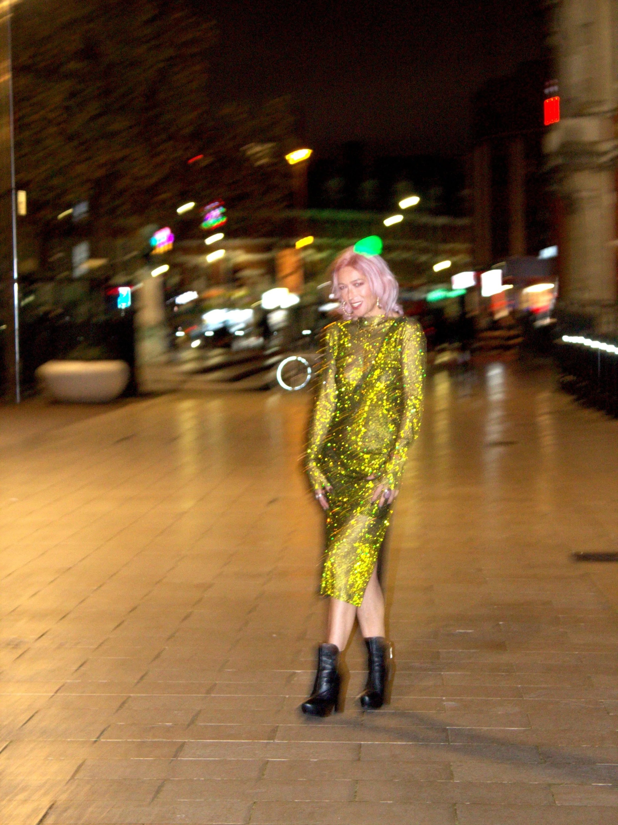 A woman with pink hair wearing a glittery yellow-green dress and black heeled boots standing on a city sidewalk at night.
