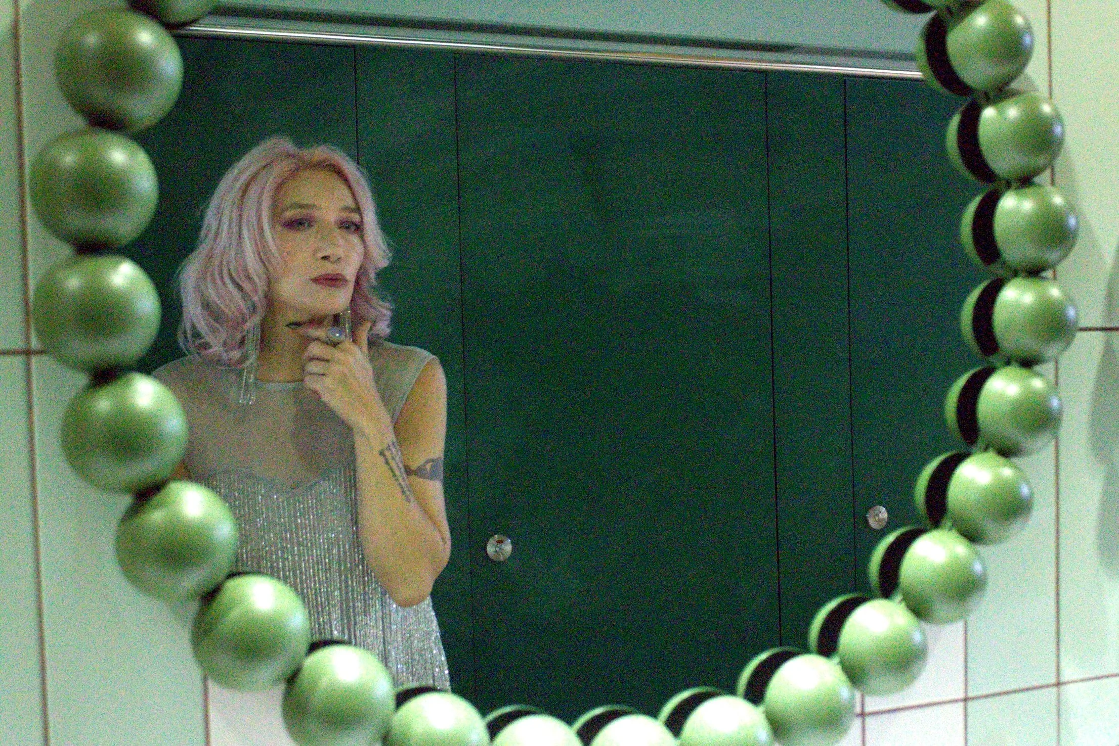 A woman with pink hair and tattoos wearing a silver dress looking into a green decorative mirror in a bathroom with green stall doors.