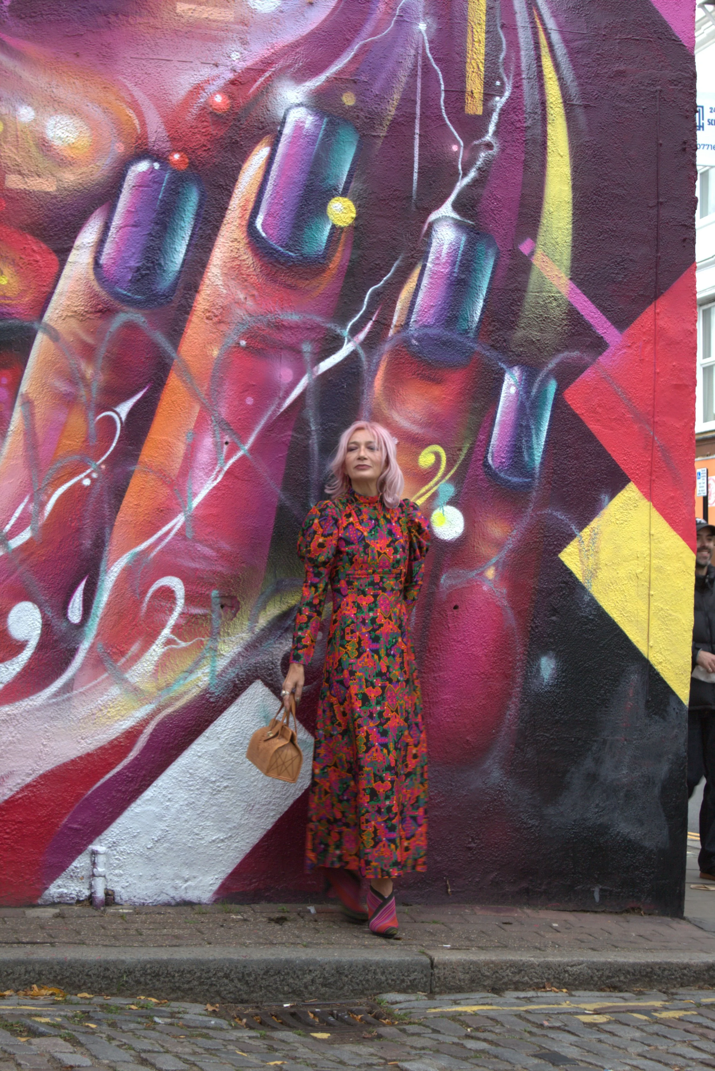 A woman with pale pink hair wearing a long colorful patterned dress and striped heels, holding a small tan handbag, standing in front of a vibrant graffiti mural of painted fingers with brightly colored nail polish, abstract lines, and shapes.