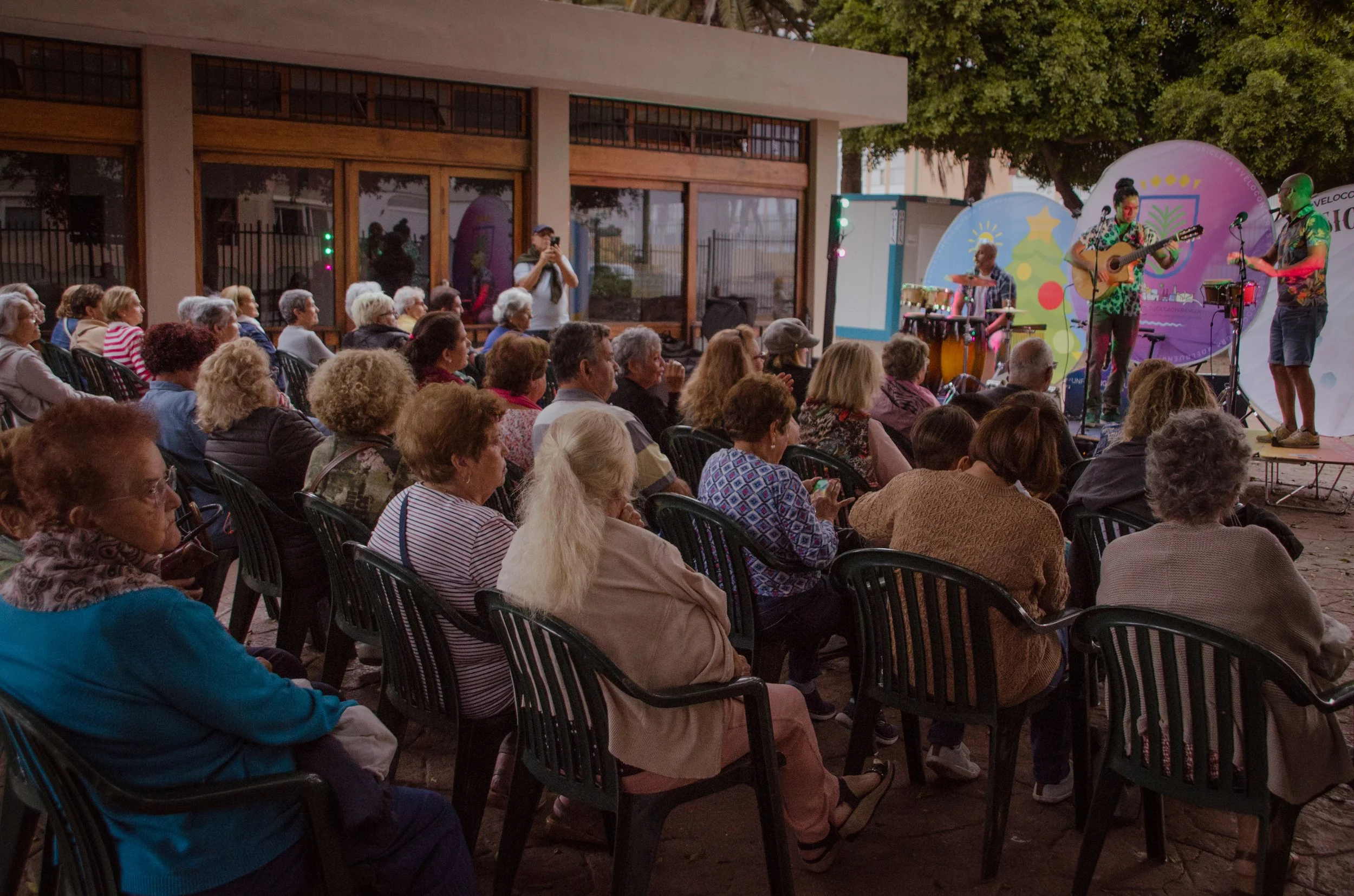 An outdoor concert with an audience of mostly elderly people sitting on black plastic chairs, watching a musical performance on stage. The stage has a colorful backdrop with holiday themes, and three musicians are playing instruments.