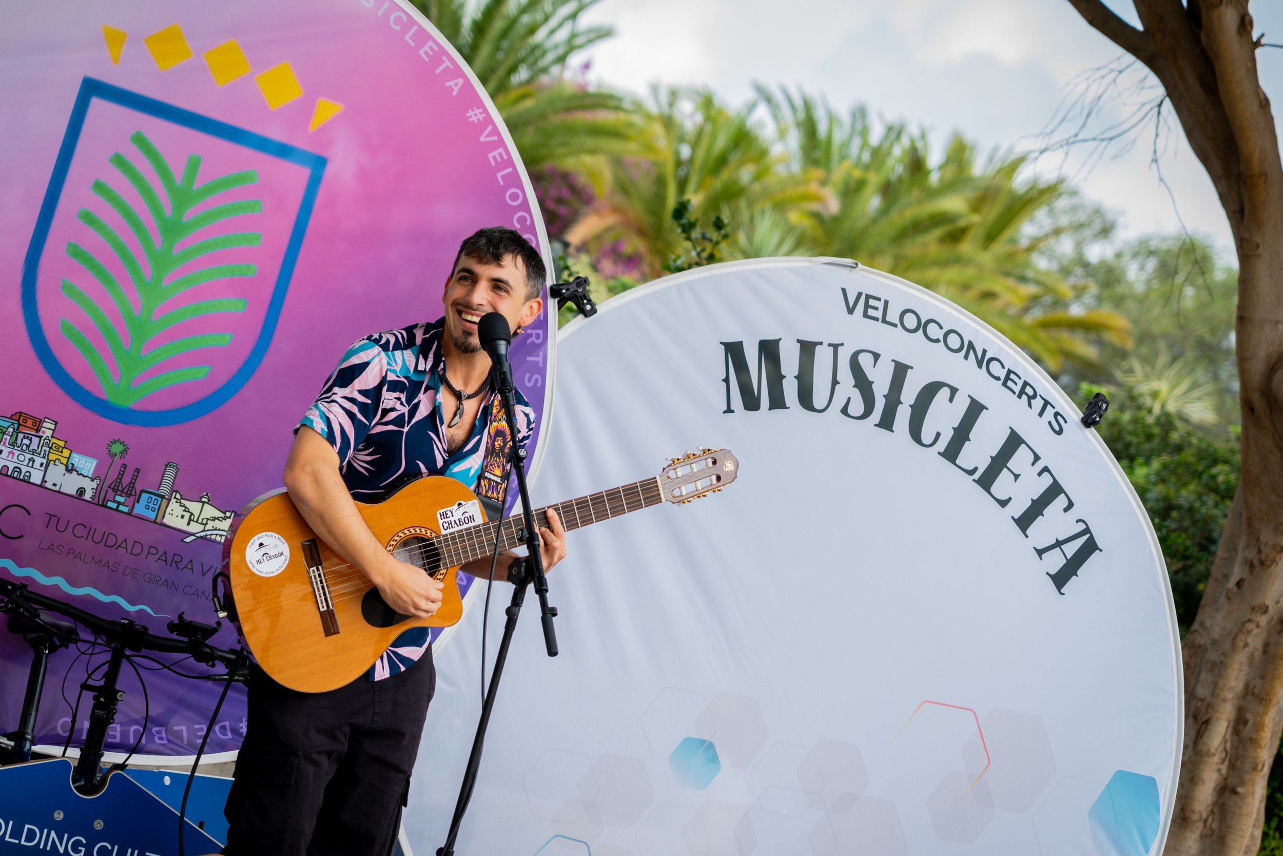 Musician performing on an outdoor stage with a guitar, smiling, with a large sign reading 'MUSICA' and greenery in the background.