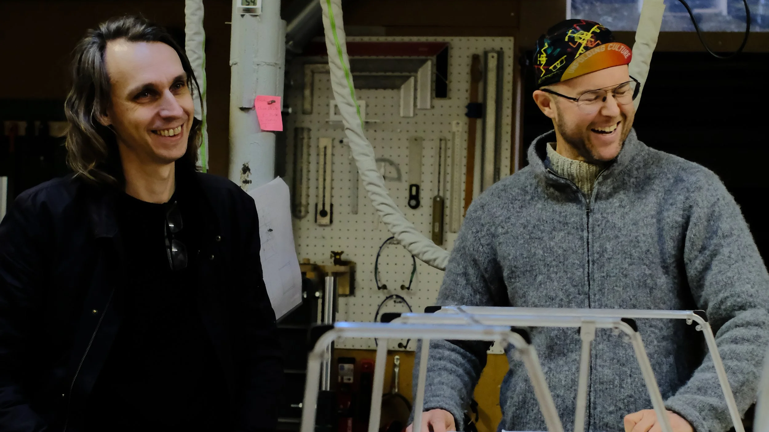 Two men smiling and laughing in a workshop or garage, with tools hanging on a pegboard behind them, and a large plastic rack or container in the foreground.