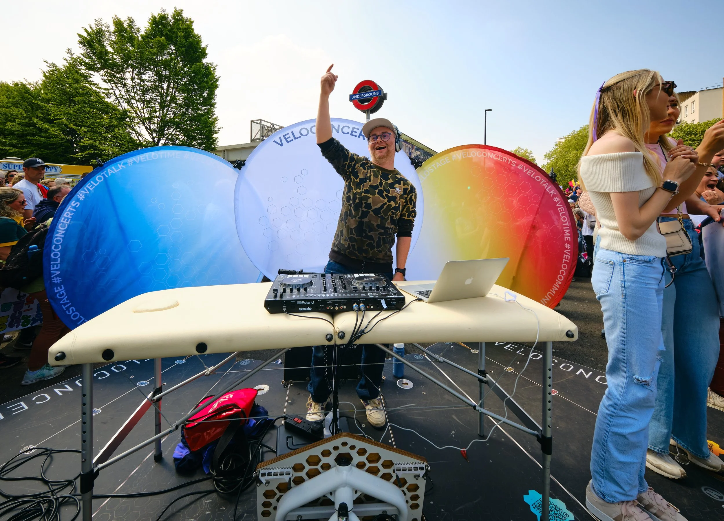 A DJ with headphones and glasses standing behind a DJ table with music equipment, smiling and pointing upward, at an outdoor event during daytime, with colorful circular decorations and a crowd in the background.