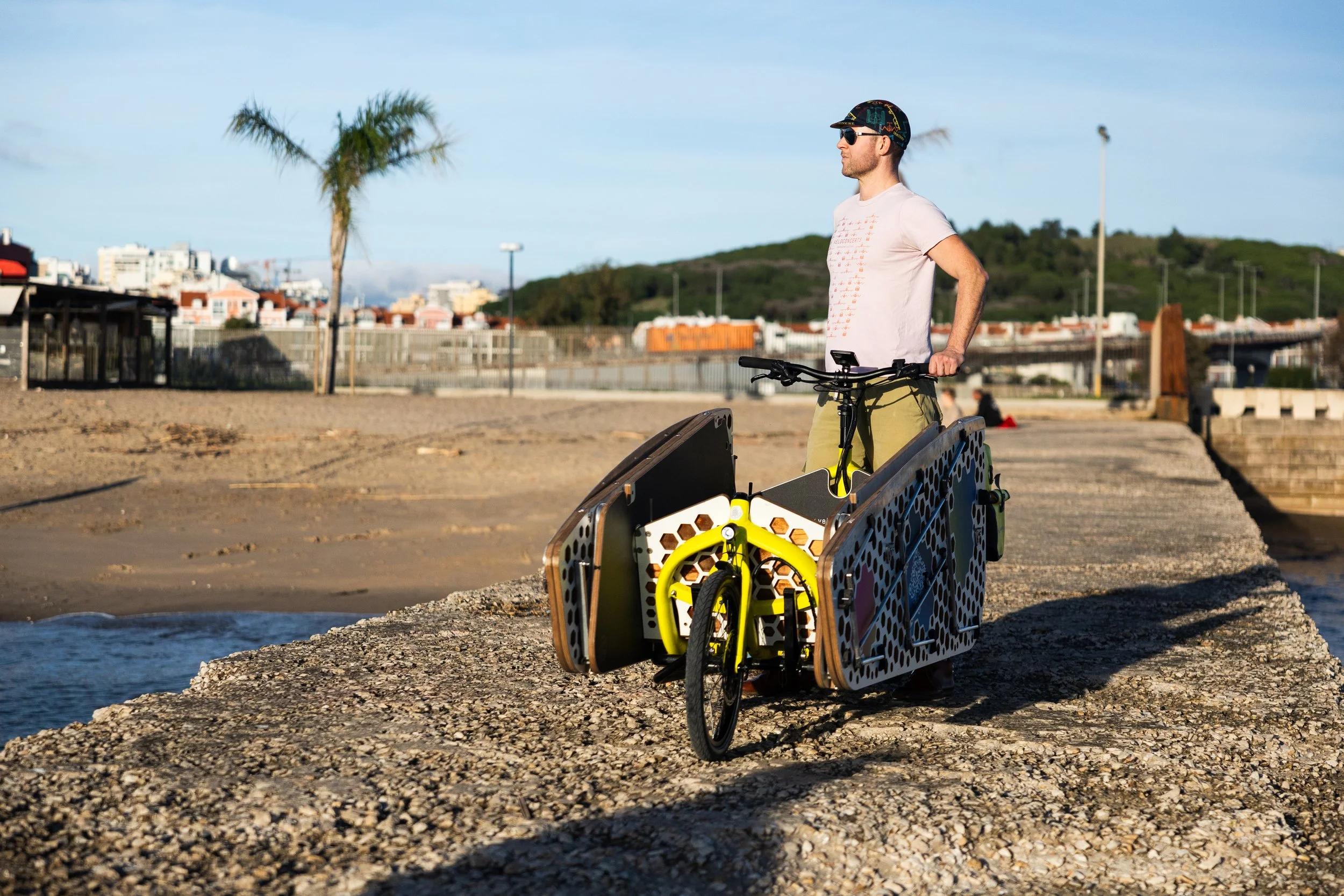 A man standing next to a bicycle with a custom trailer attached, near a beach with a pier, sandy shore, palm trees, and a cityscape in the background.