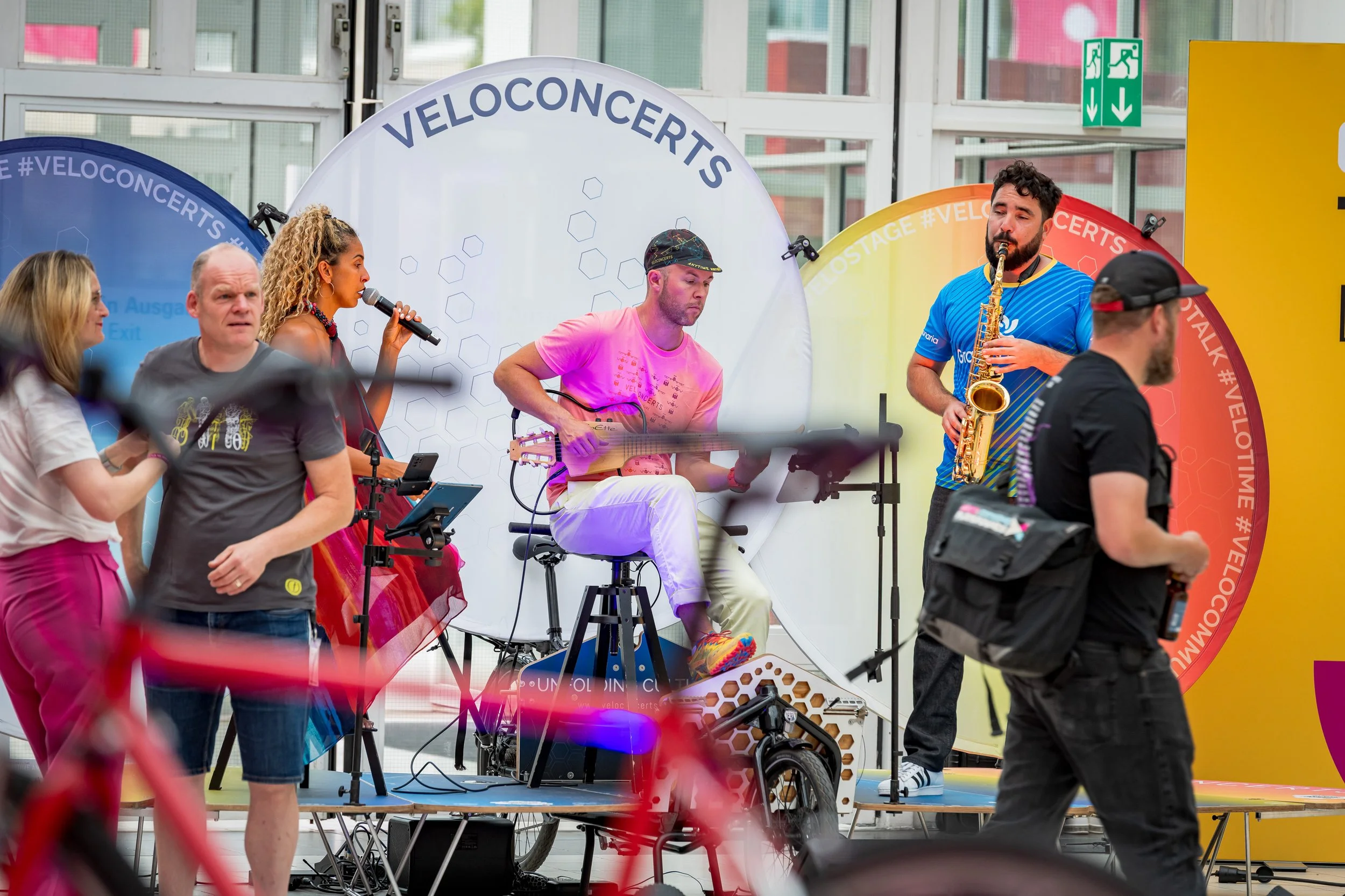 Musicians performing on a stage at a public event, with a woman singing into a microphone, a man playing a keyboard, and another man playing a saxophone. The stage features colorful circular backdrops with the hashtag #VELOCIGHTS.