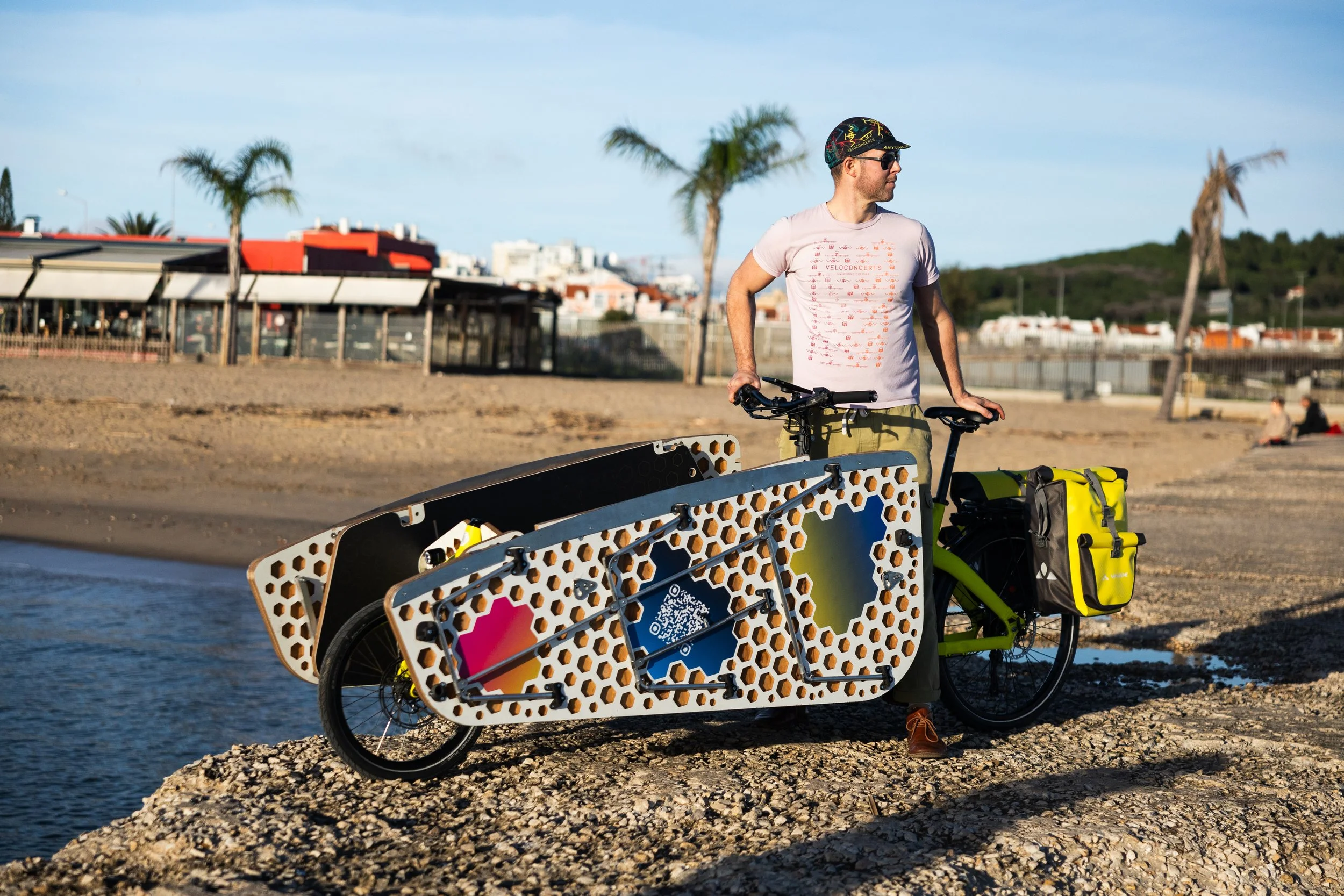 A man standing beside an orange and black cargo bicycle with a large, flat, honeycomb-patterned platform, on a rocky beach near water. He wears a white t-shirt, khaki pants, sunglasses, and a black cap. In the background, there are palm trees, a building with a red and white roof, and a clear, blue sky.