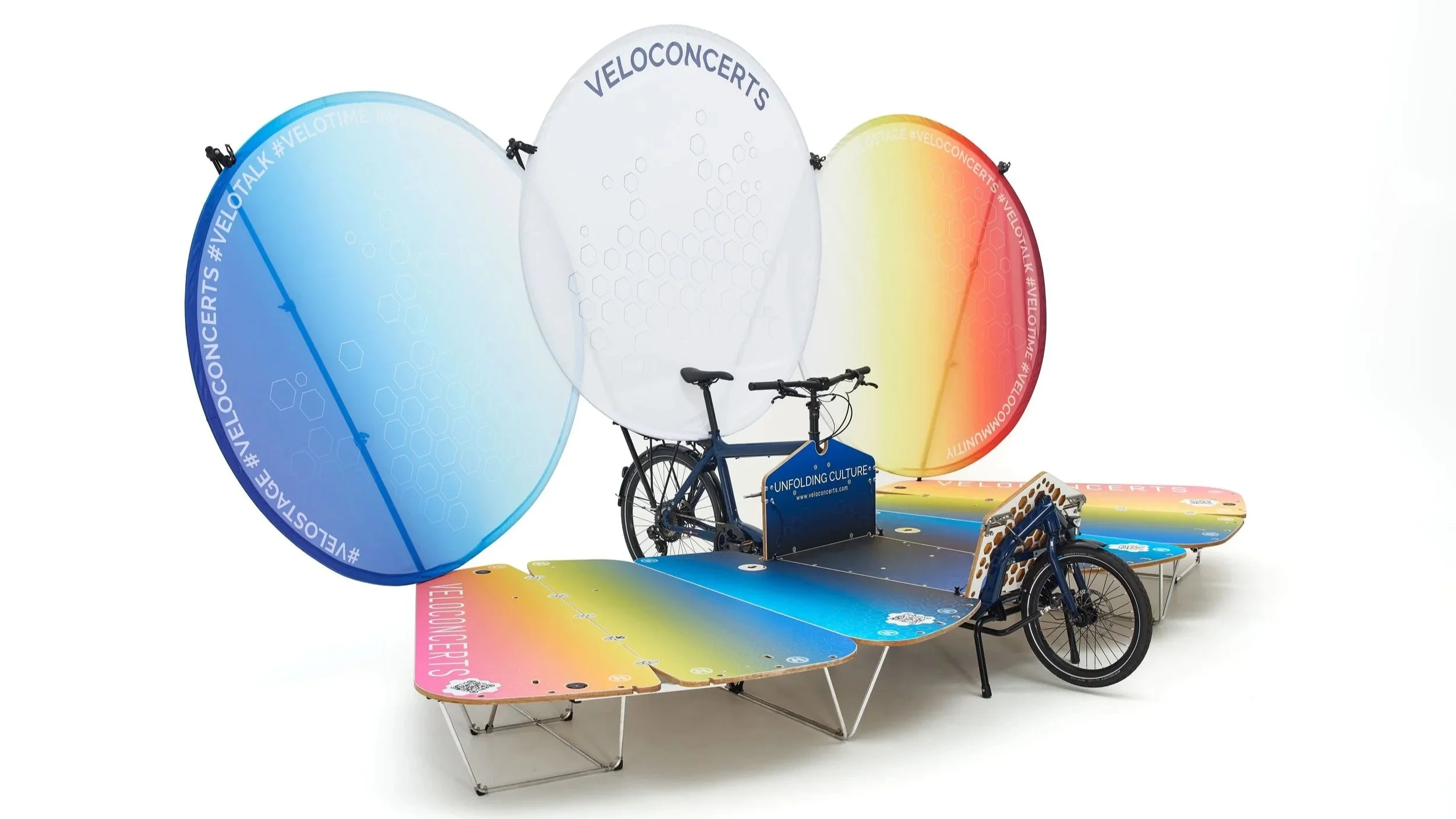 A colorful mobile outdoor display featuring a bicycle attached to a platform with large pastel-colored, circular windshields in blue, white, and yellow, decorated with hashtags and the words 'VELOCONCERTS', promoting an event or campaign.