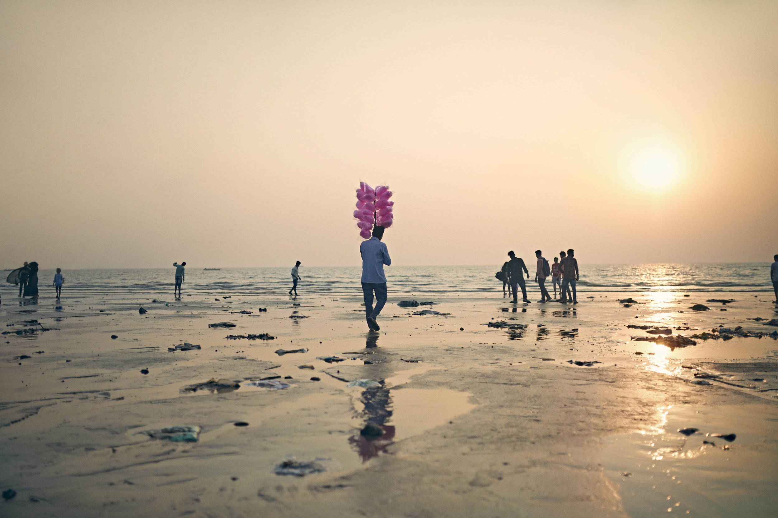 A man holding a large tower of candy floss (cotton candy) walks down an beach in Mumbai, India. By lifestyle photographer Jon Enoch