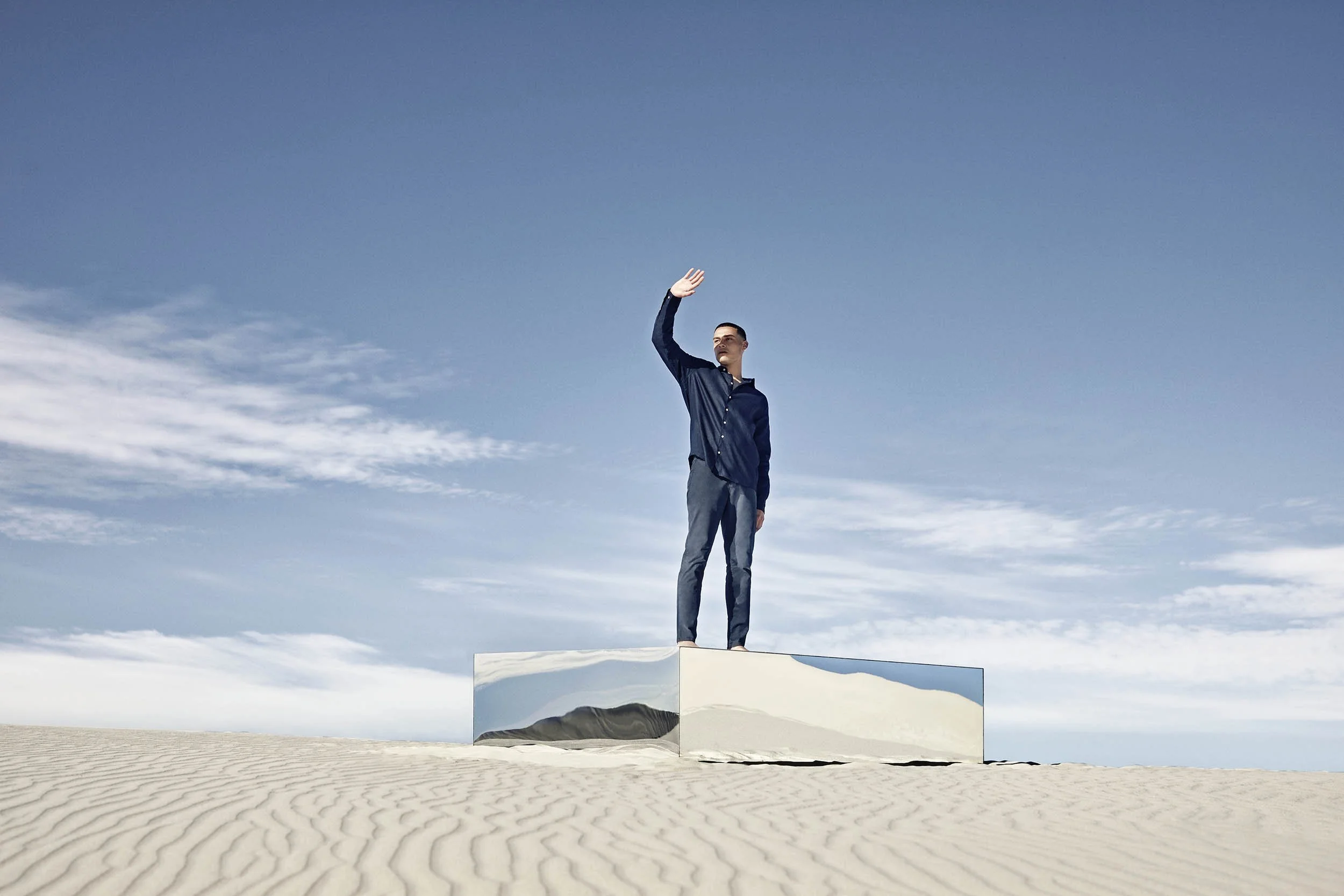 A man wearing a blue shirt stands on-top of a mirror in the desert in this conceptual image from lifestyle photographer Jon Enoch