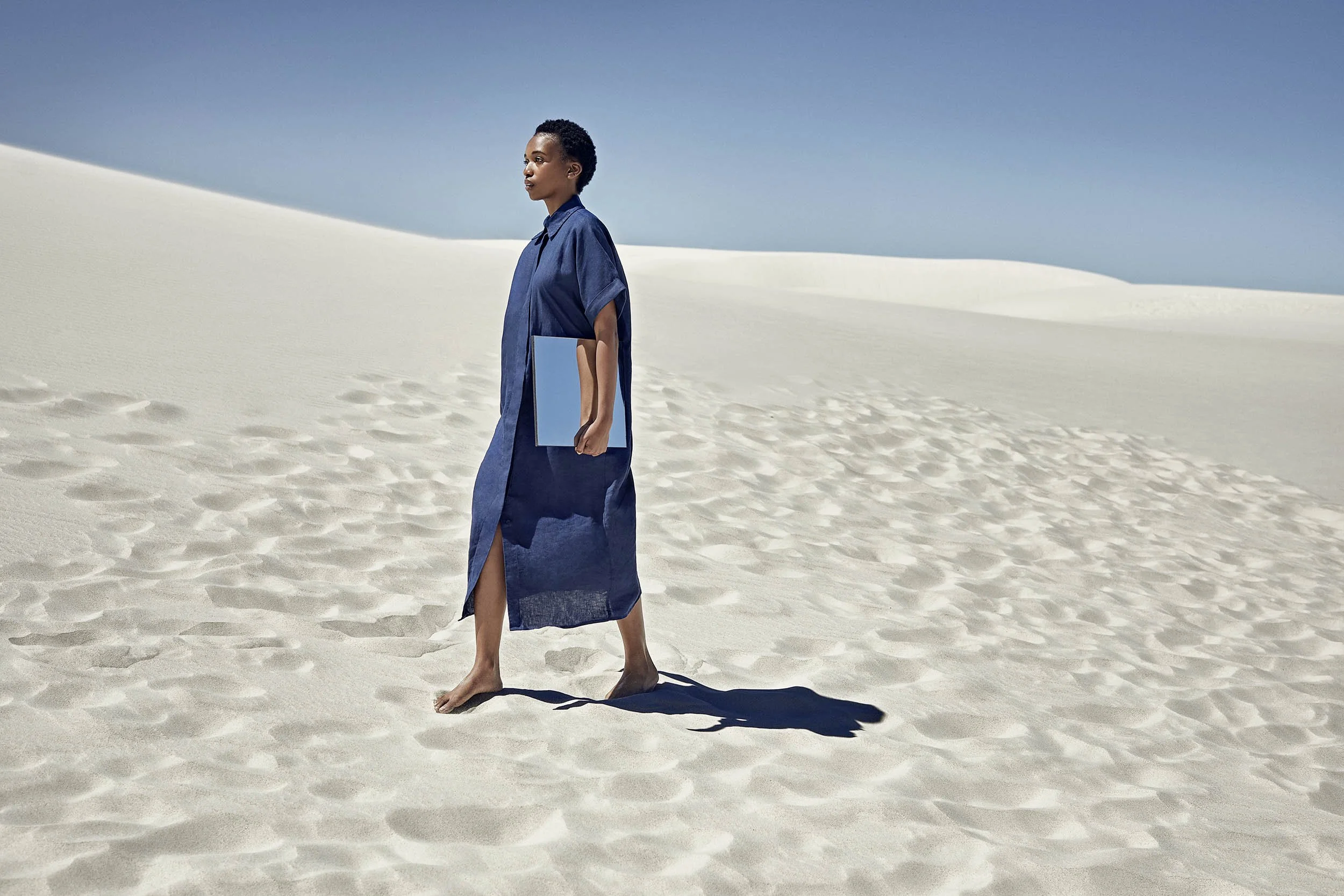 A women wearing a blue dress walks barefoot across sand holding a mirror under her arm in this advertising lifestyle image by photographer Jon Enoch
