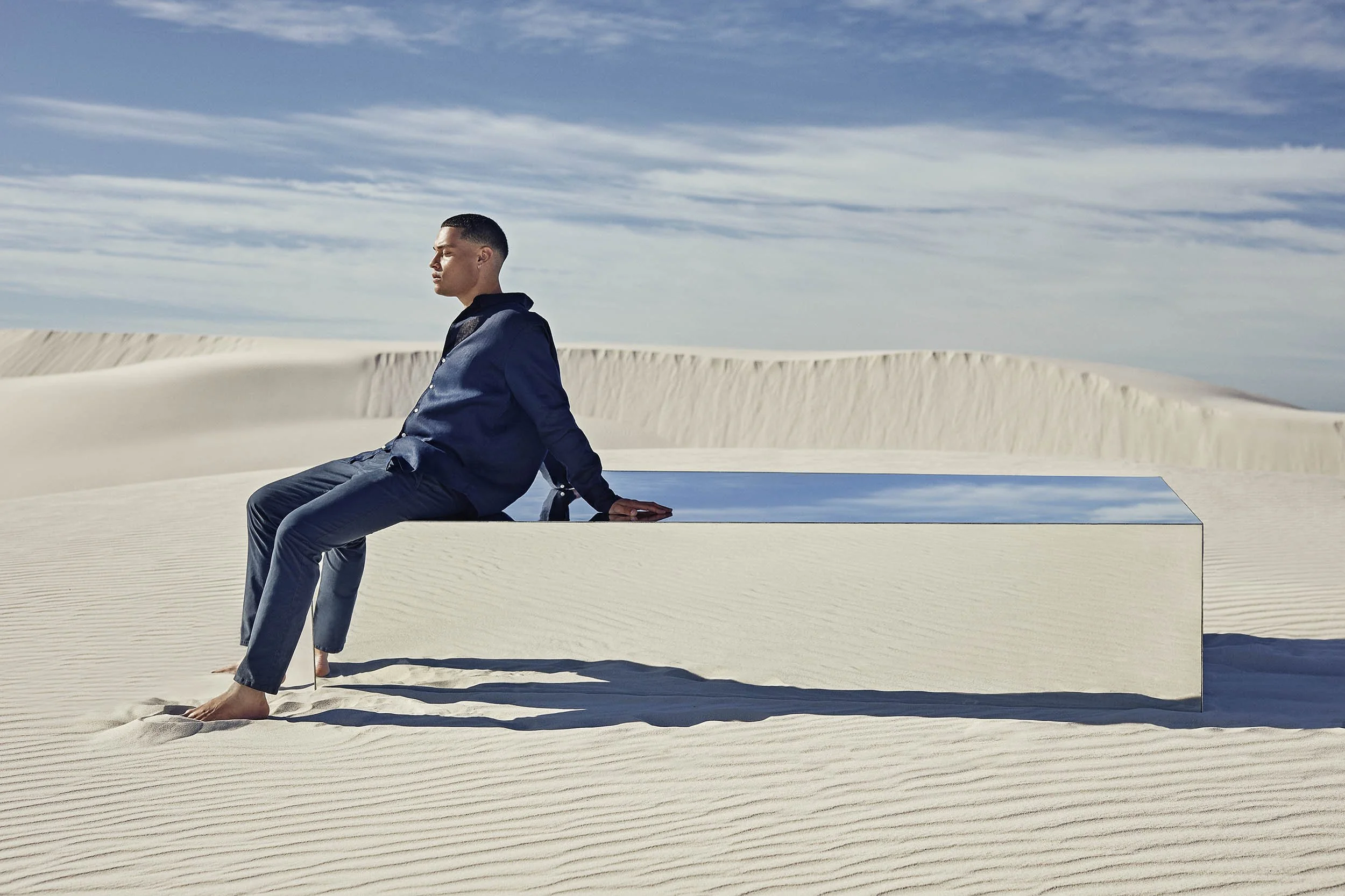 A man sits on a large mirrored box in the desert the mirror reflecting the sky in this lifestyle image by Jon Enoch