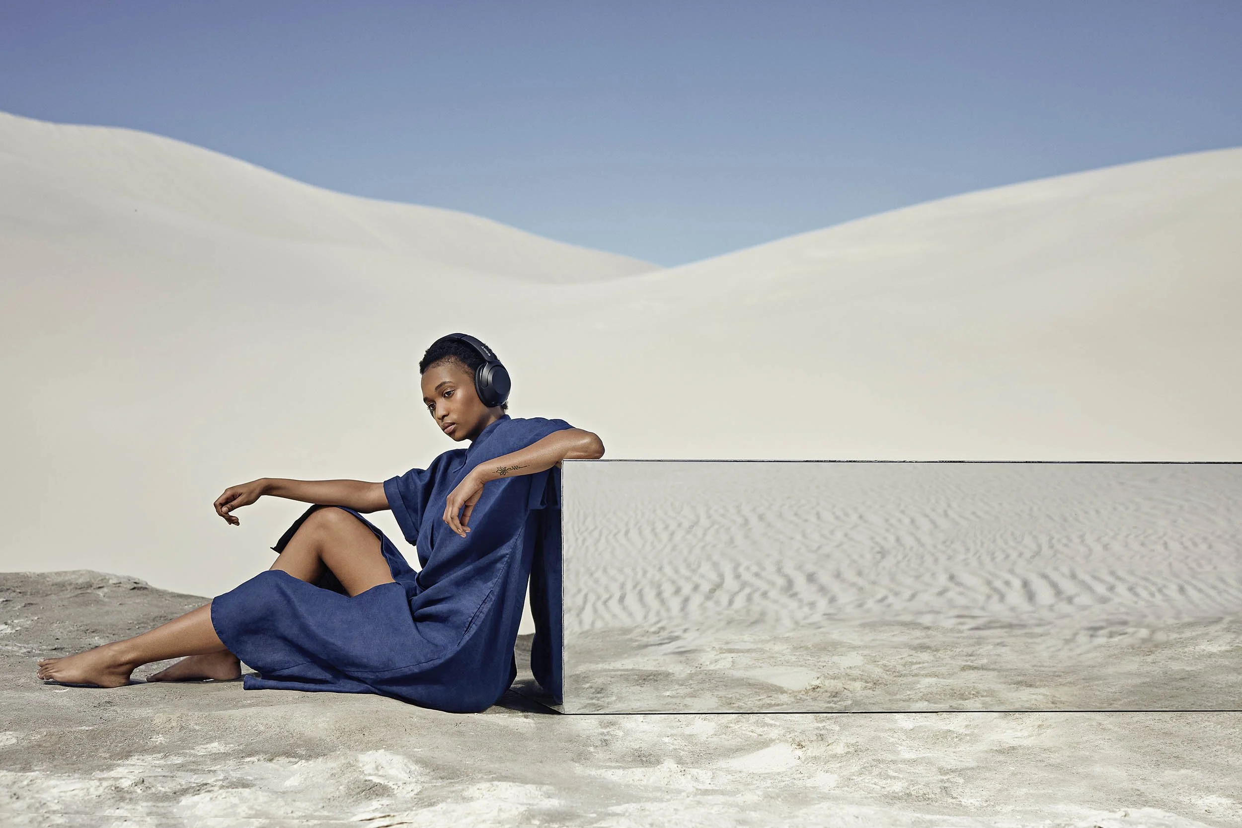 A women listens to music on headphones whilst sitting next to a large mirror in the desert in this lifestyle image by Jon Enoch
