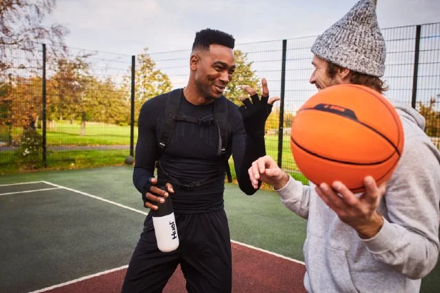 Two men, one holding a Huel shaker chat on a basketball court.