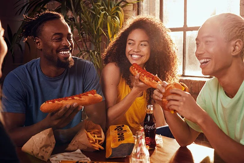 A group of friends eat hotdogs, burgers and chips whilst having a good time. Lifestyle photography by Jon Enoch.