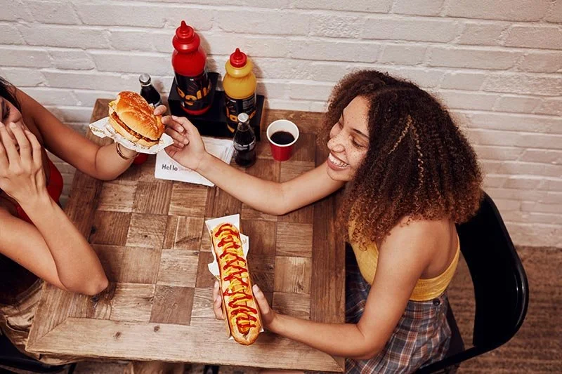 Two friends laugh and chat whilst eating food and drinking drinks in the lifestyle photography by Jon Enoch.