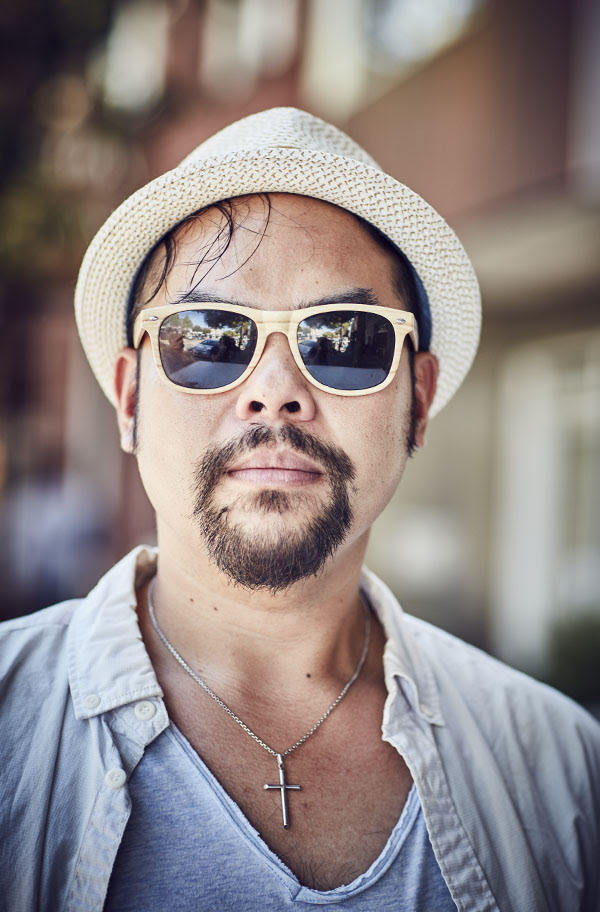 A man wearing a hat and dark glasses poses in the street. In the Mission, San Francisco. Photography by portrait photographer Jon Enoch.