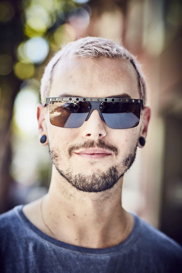 A man wearing futuristic glasses poses in the street. In the Mission, San Francisco. Photography by portrait photographer Jon Enoch.