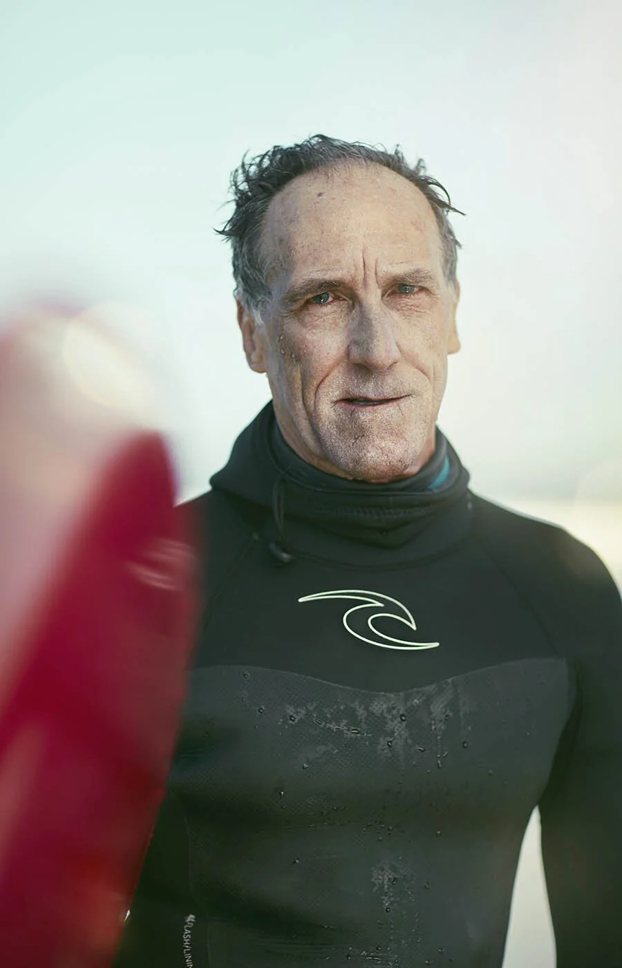 A older male surfer poses for a picture whilst holding his long board on the beach in California in this photography taken by London based sports advertising photographer Jon Enoch.