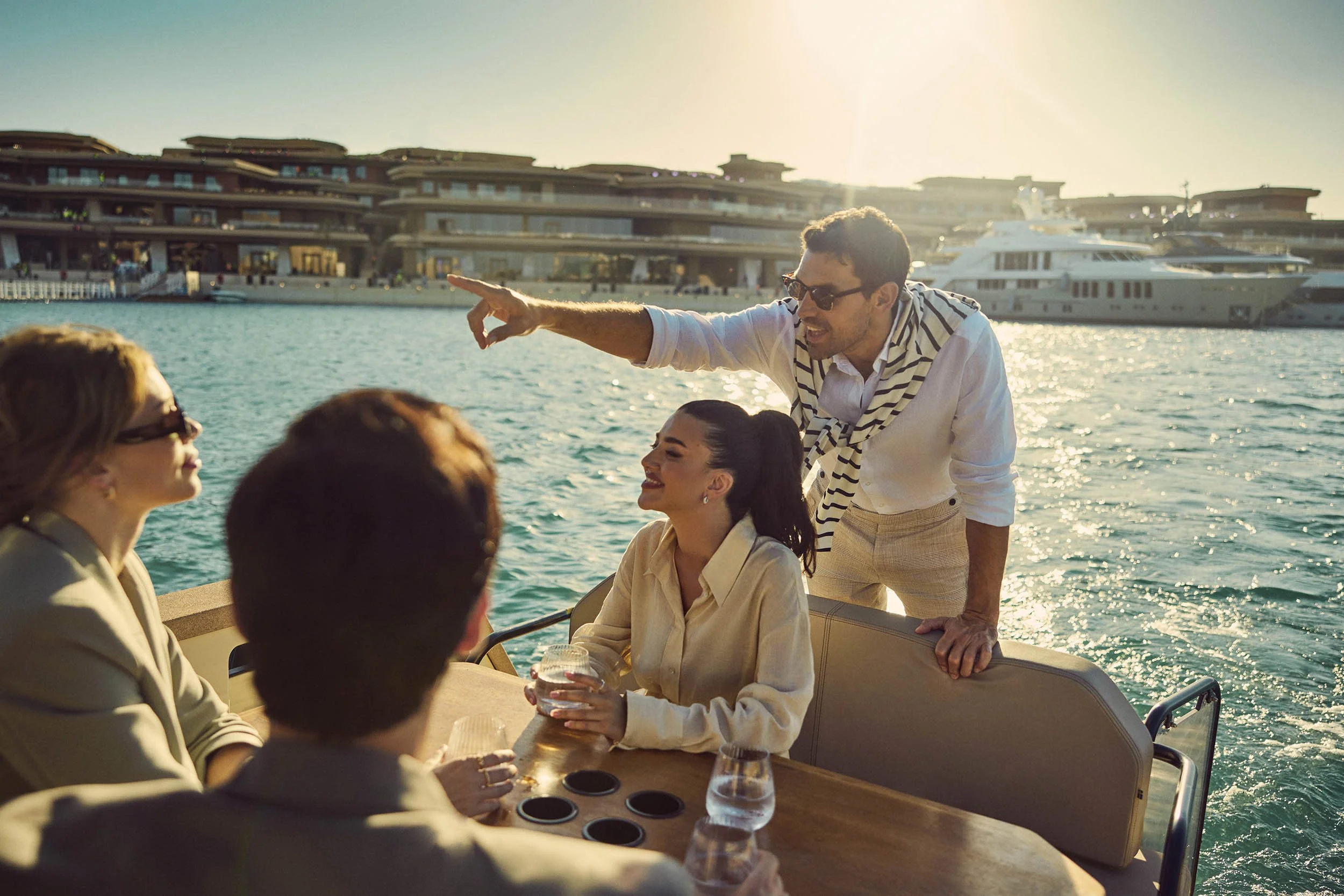 People enjoying a boat ride near yachts, with buildings in the background, on a sunny day by the water.