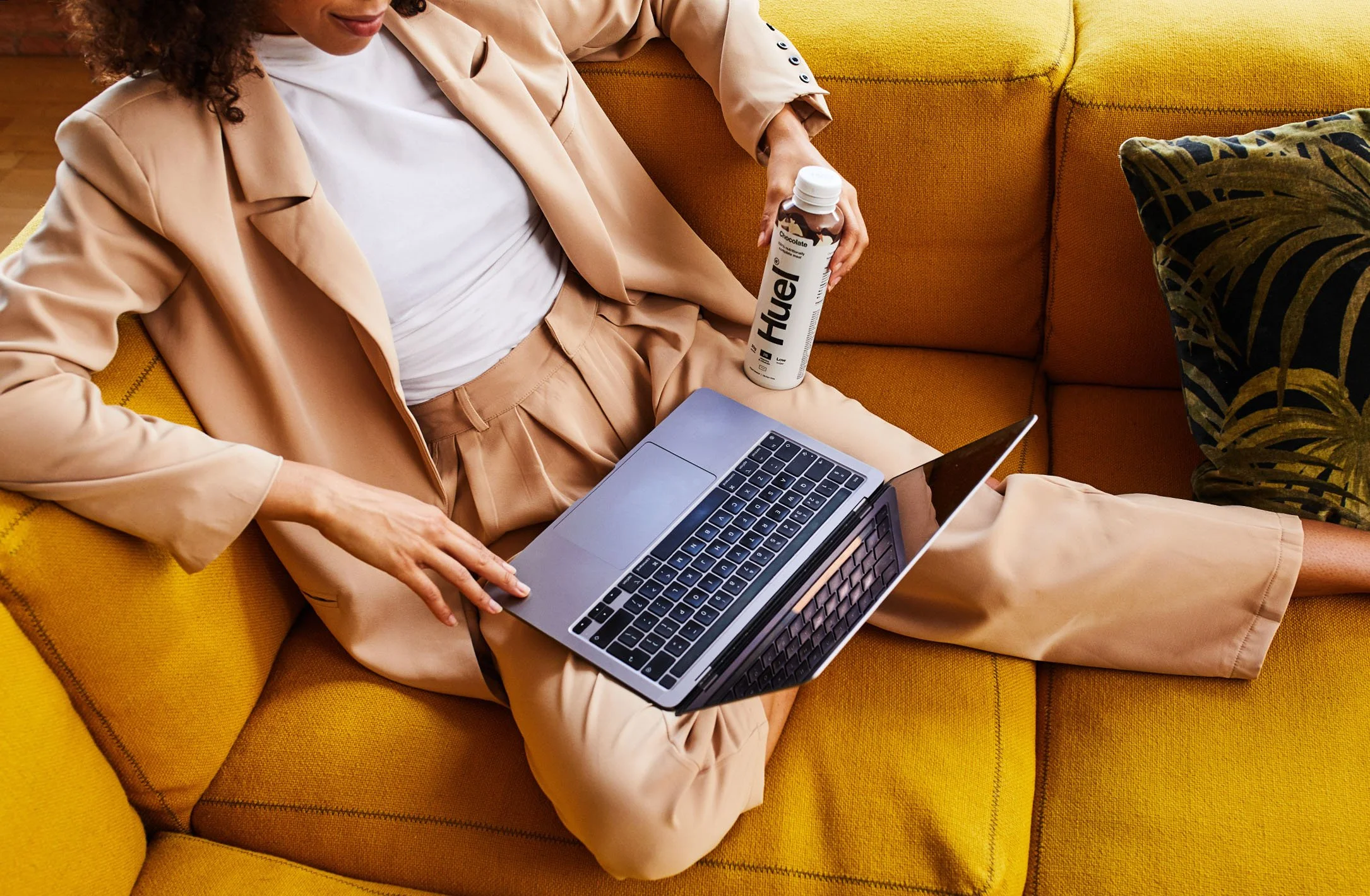 A woman in beige business attire sitting on a yellow couch, using a laptop with her feet up, holding a bottle of Huer natural energy drink.