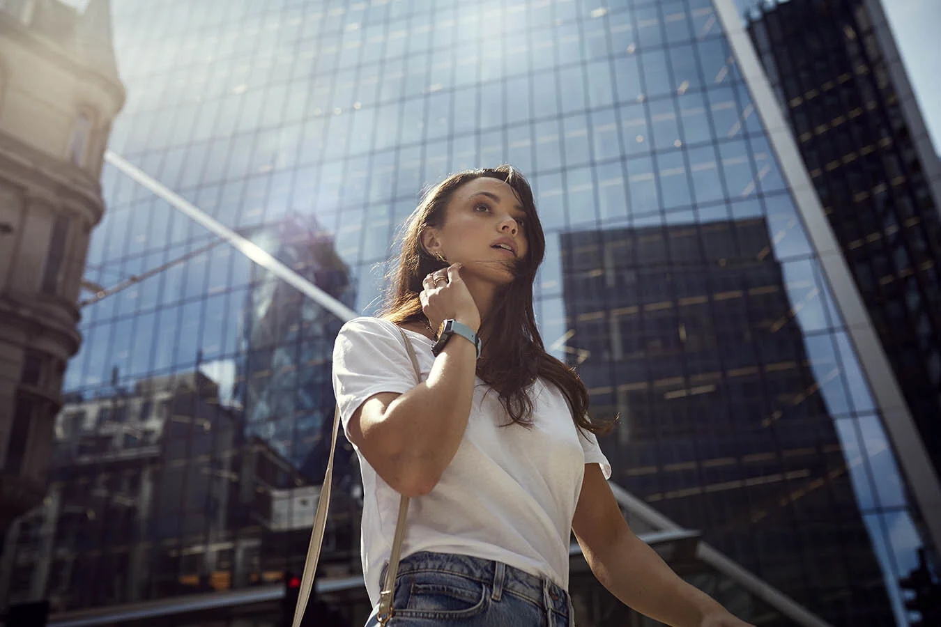 Young woman with long brown hair wearing a white t-shirt and jeans, standing outdoors in an urban area with tall glass buildings reflecting the sky and other buildings, sunlight shining on her.