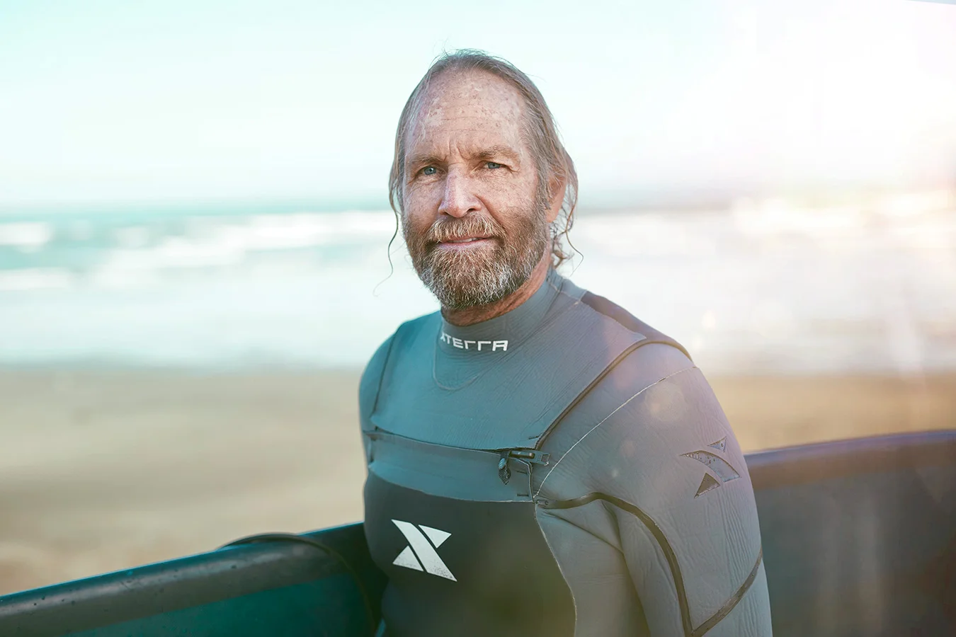Middle-aged man with gray hair and beard wearing a wetsuit, holding a surfboard on a beach with ocean waves in the background.