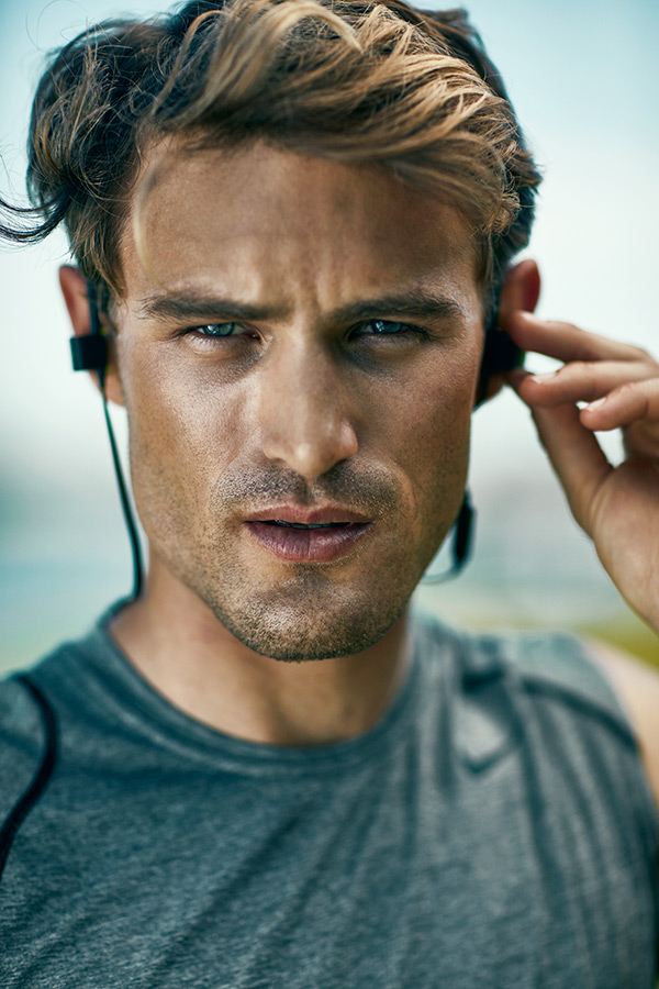 Close-up of a young man with tousled hair, wearing a gray athletic shirt, adjusting his headphones with a focused expression.