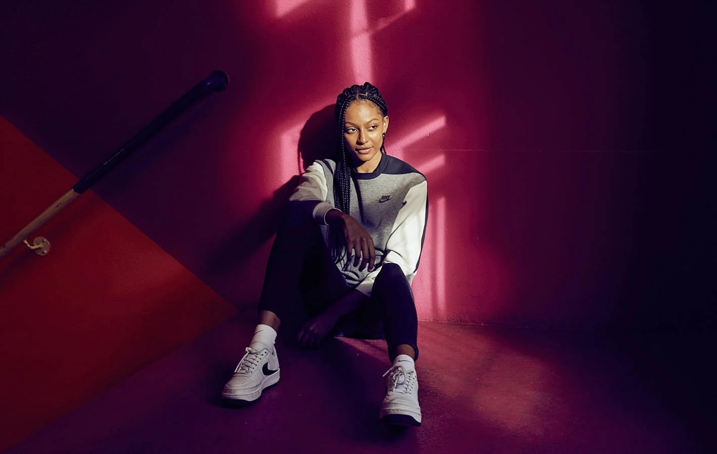 A young woman with braided hair sitting on the floor against a red and purple wall, wearing a Nike sweatshirt, black pants, and white Nike sneakers.