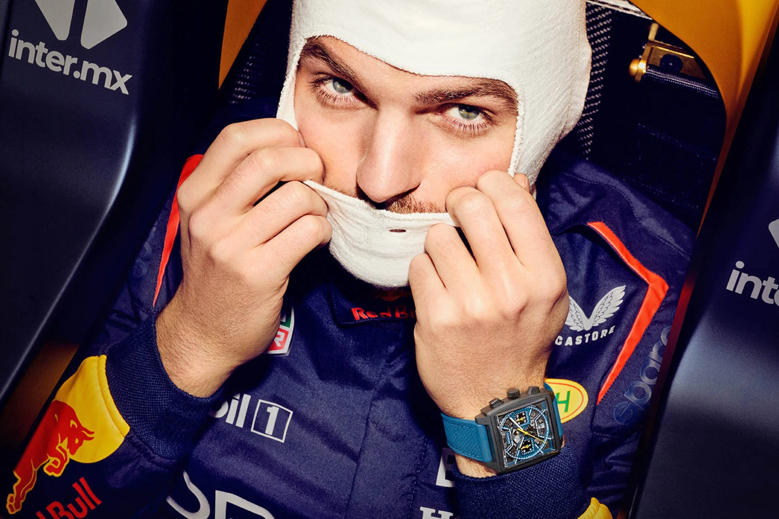 A man in a racing suit with Red Bull and other sponsor logos is pulling a white headband or cap over his head, revealing his face and eyes.
