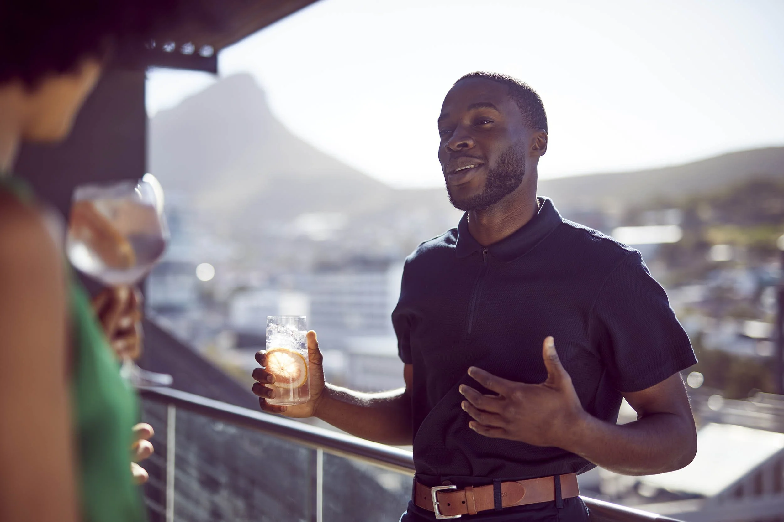 A man chats to his friends, drink in hand on a rooftop in summer in this lifestyle drinks shot by Jon Enoch
