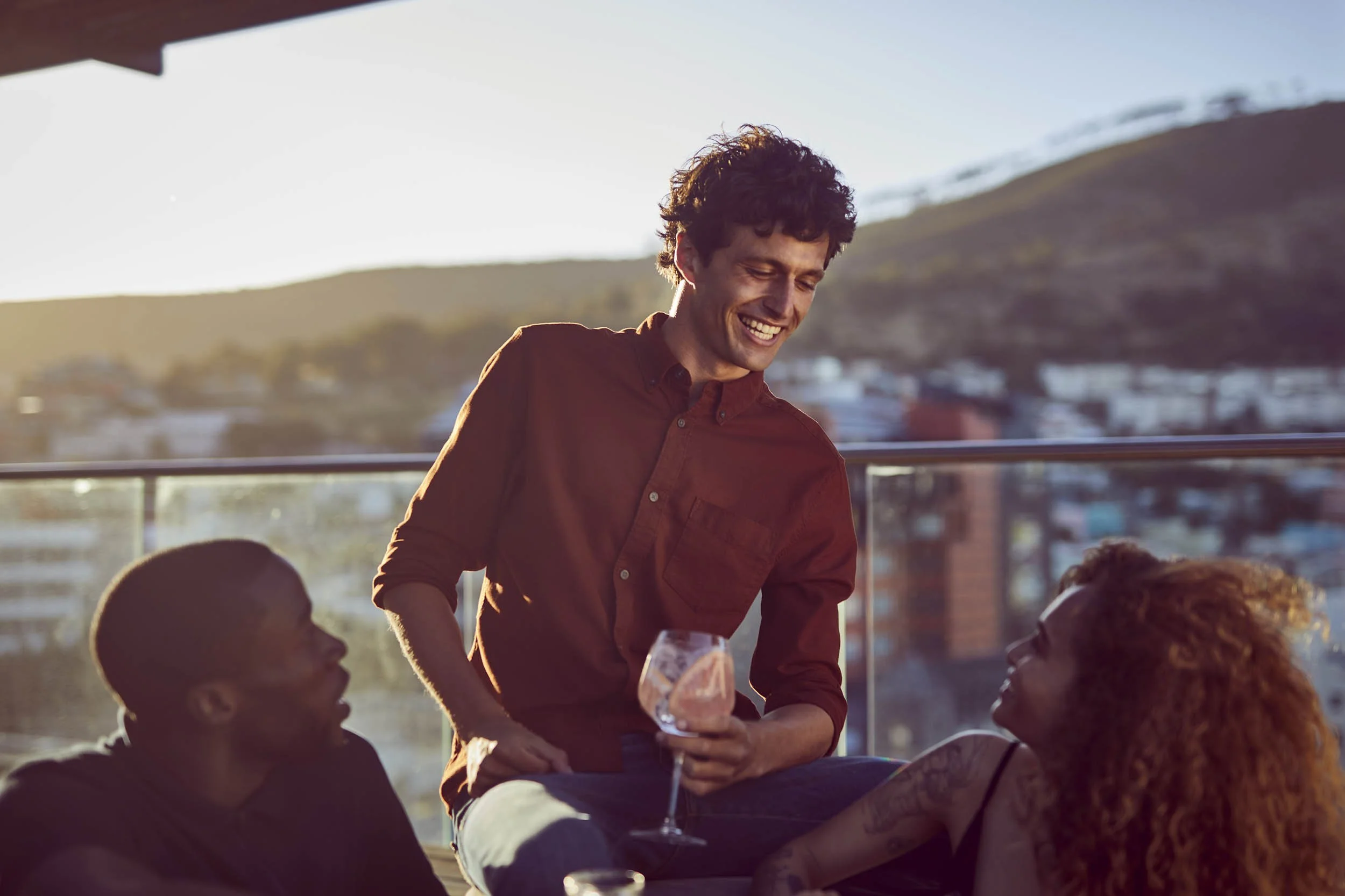 A young man chats to his friends as they sit on a rooftop and watch the sun set whilst drinking a few summer drinks - lifestyle image by Jon Enoch