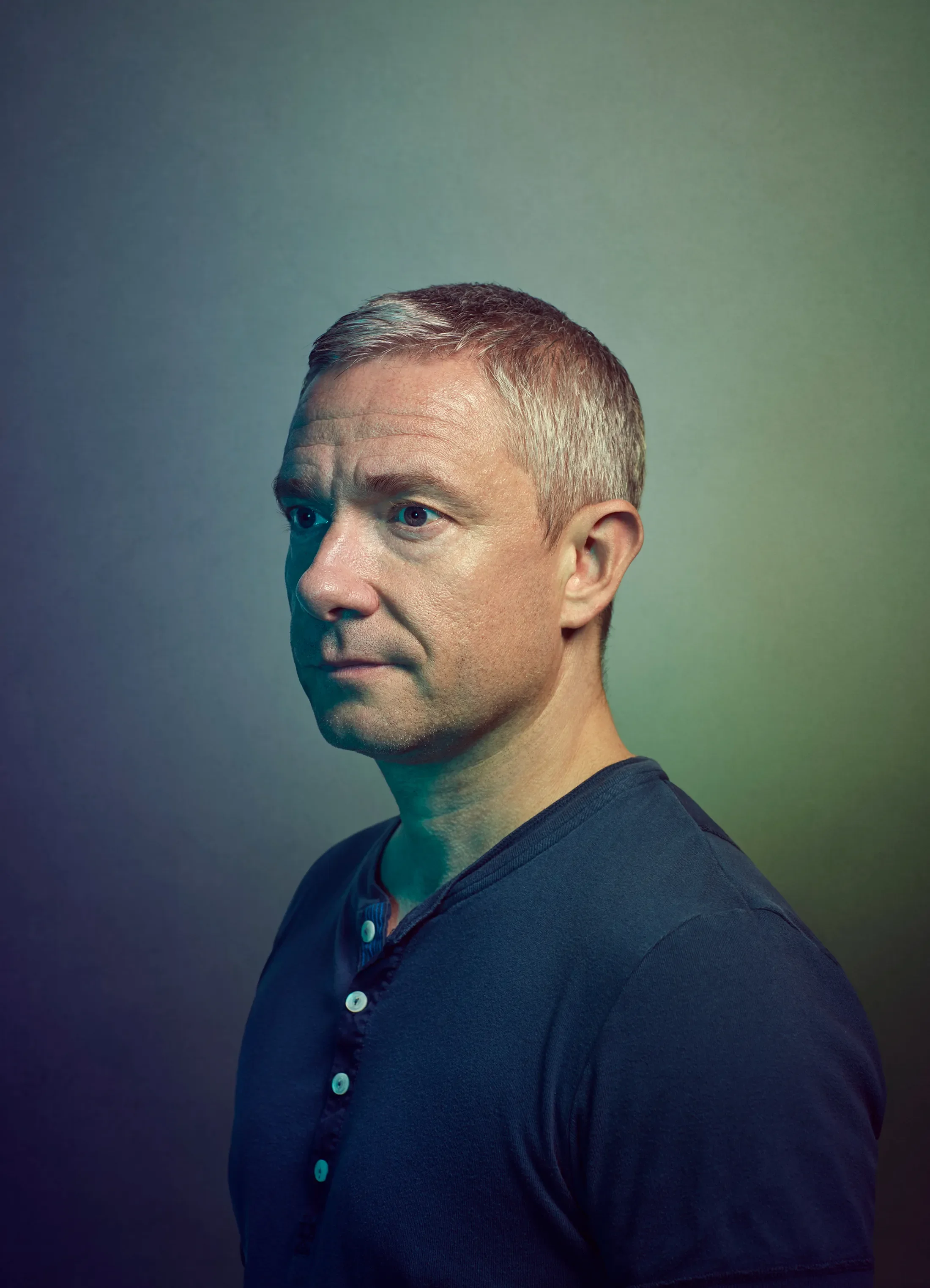 A middle-aged man with short, gray hair wearing a dark blue shirt, looking slightly to the side against a gradient background.