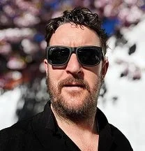 Man with dark curly hair and beard wearing black sunglasses and a black shirt, standing outdoors with blurred trees and sky in the background.