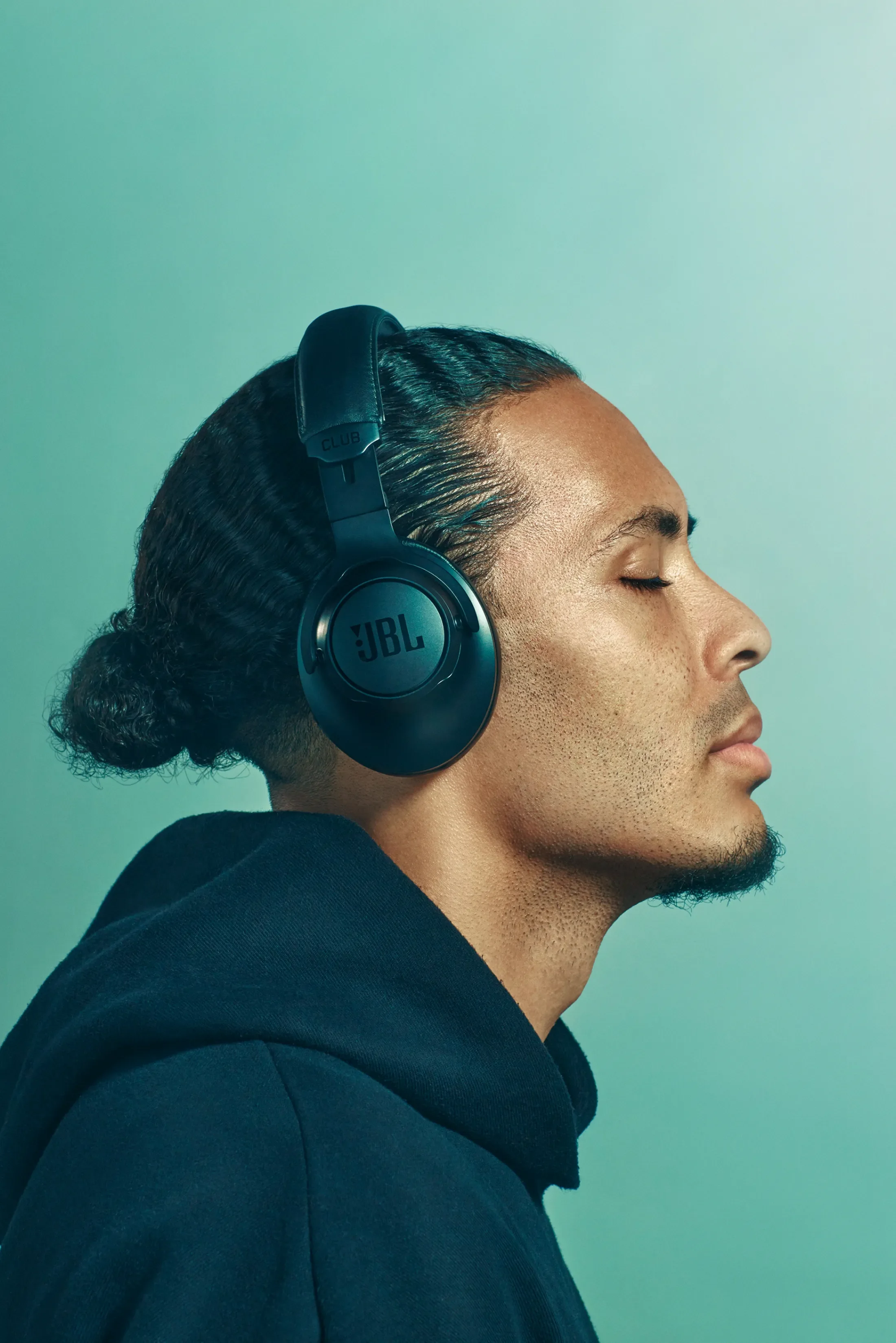 A man with short, slicked-back hair and a beard, wearing a black hoodie, is listening to music with black JBL headphones, with his eyes closed.