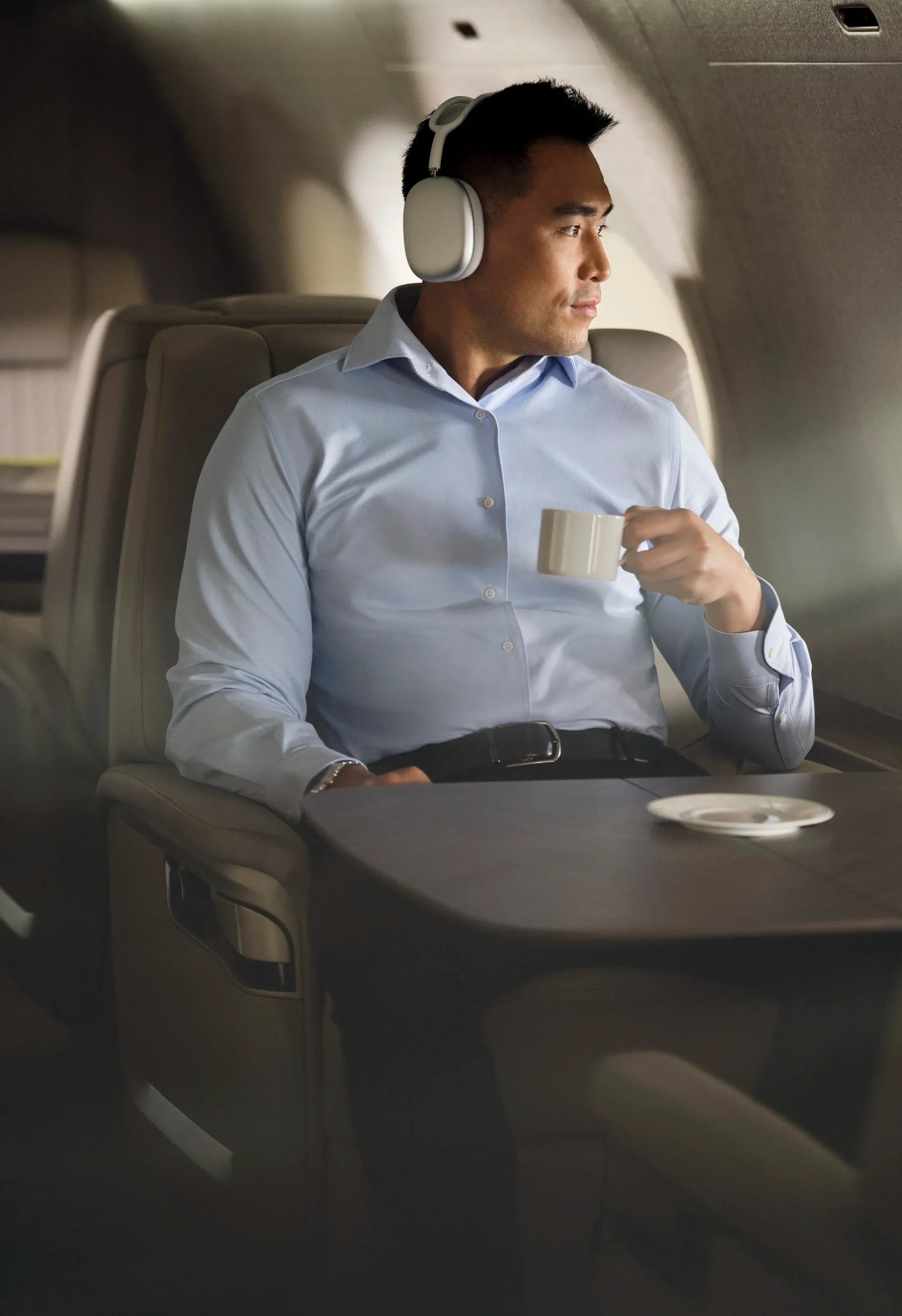 Man in a light blue shirt with headphones, sitting in an airplane seat, holding a coffee cup, and looking out the window.