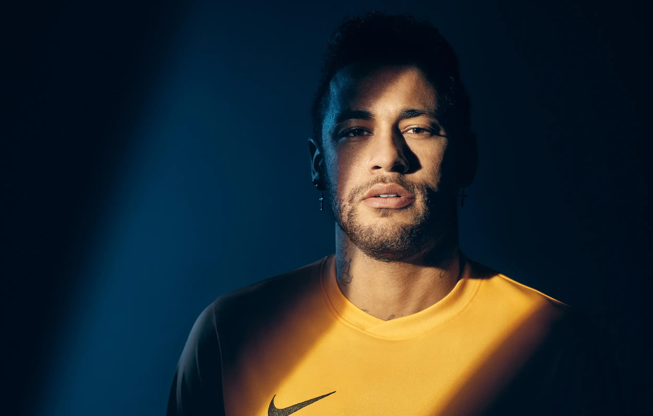 A young man with dark hair and earrings, wearing a yellow Nike sports shirt, looks directly at the camera against a dark background with a spotlight across his face.