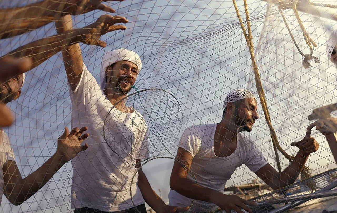 A group of men wearing white shirts and traditional Yemeni headscarves, standing behind a fishing net on a boat, engaging in fishing activities.