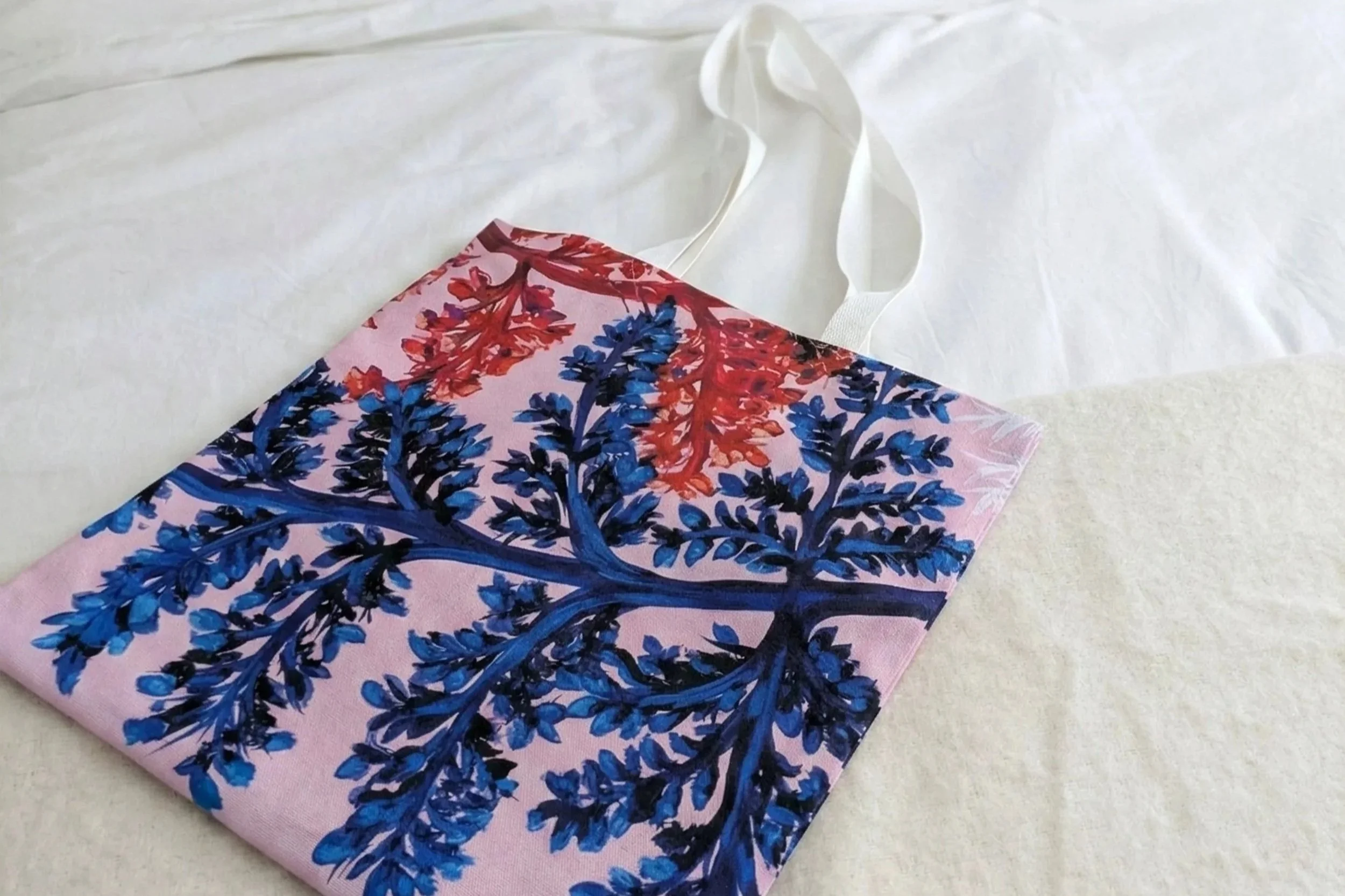 Close-up of a custom 100% cotton tote bag resting on white bedding, highlighting the vibrant blue and red print and durable fabric. A bespoke product for artists from OH Arts.