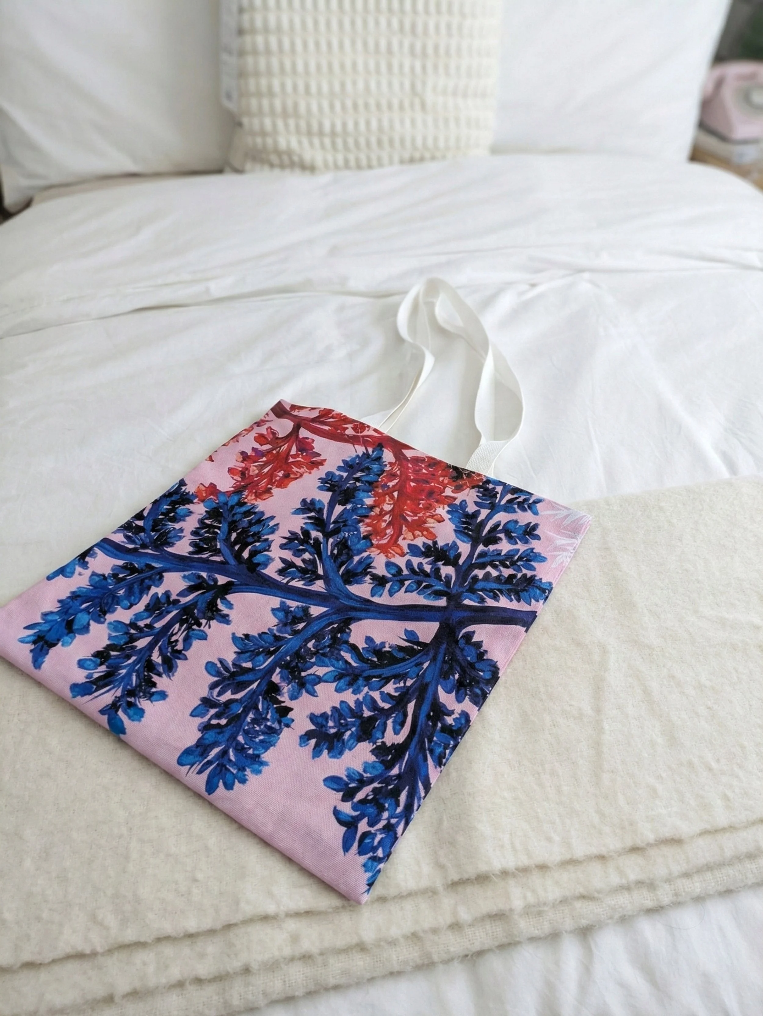 A vibrant custom canvas tote bag resting on a cream blanket at the foot of a white-sheeted bed. It showcases the bold blue and red fern foliage pattern by OH Arts. Rigorously tested quality merch for creatives.