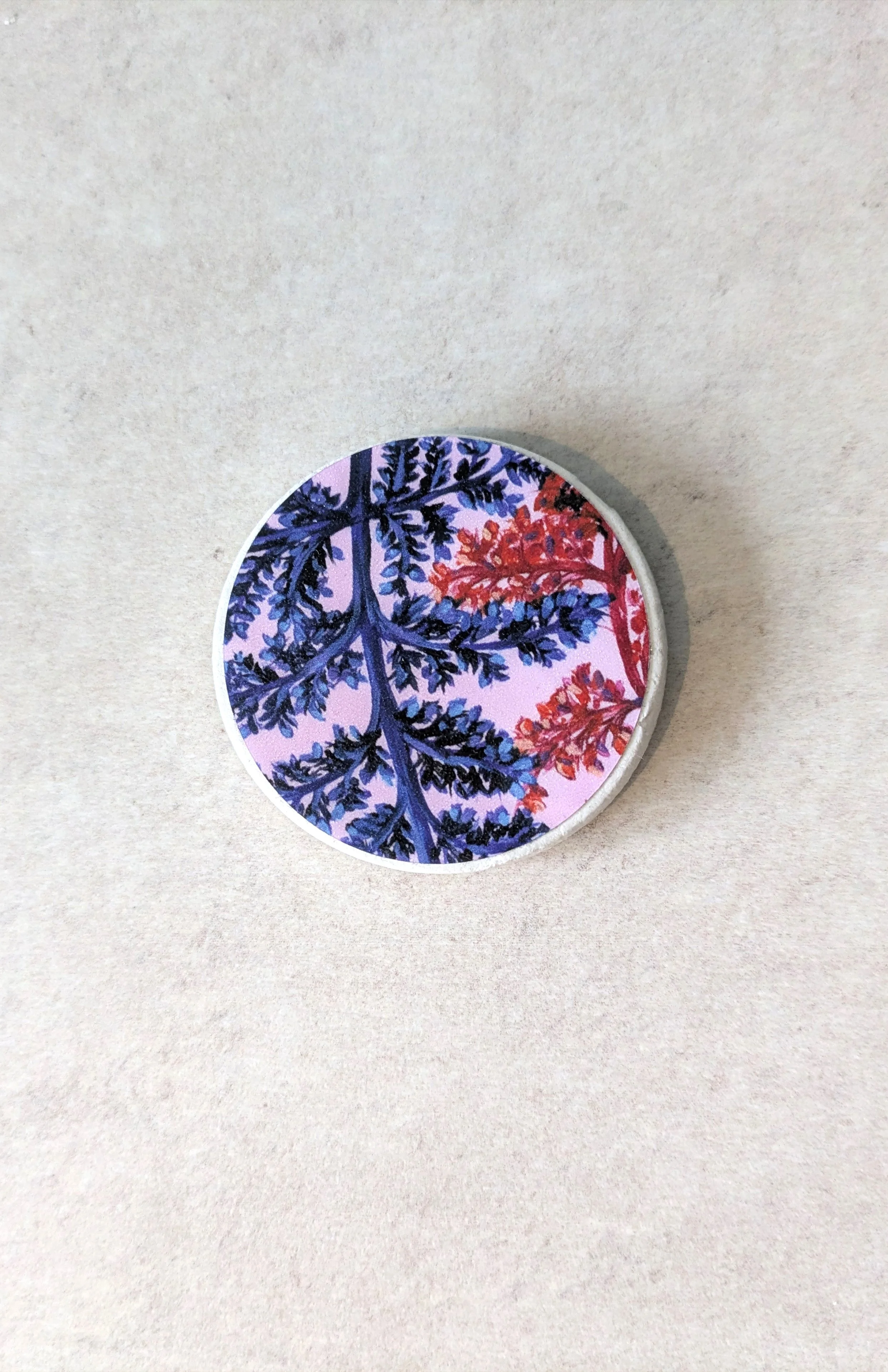 A single, round handmade clay magnet by OH Arts, showcasing a vibrant blue and red botanical illustration design on a textured background.