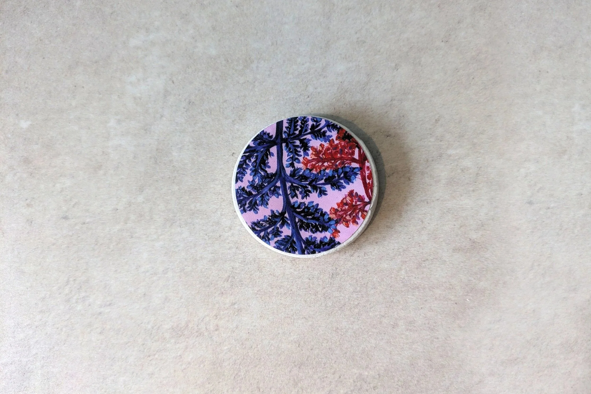 A close-up photograph of a single, handmade round clay magnet by OH Arts. It features an intricate blue and red botanical fern illustration on a soft pink background. Premium custom giftware for artists.